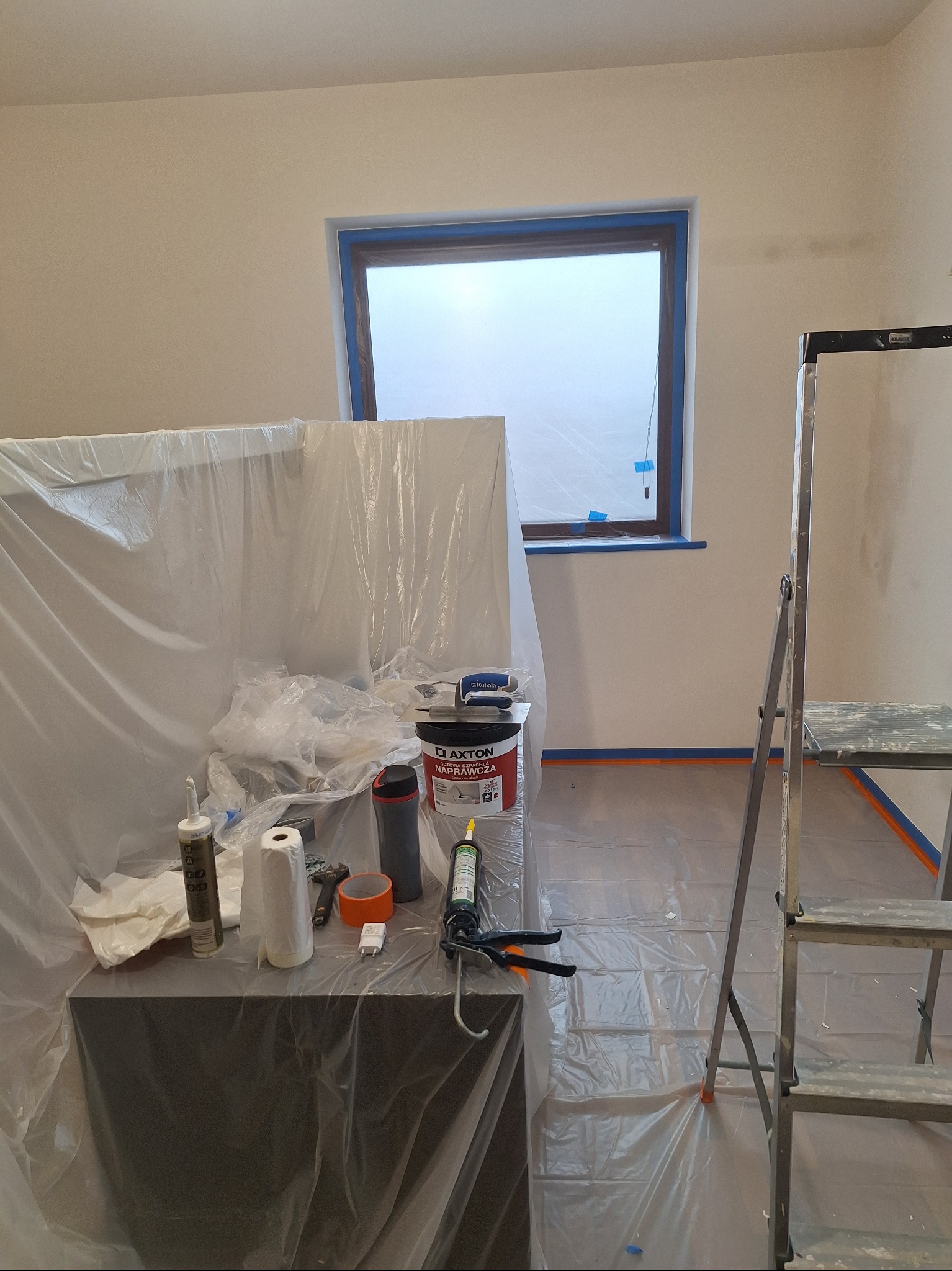 Przygotowania do malowania: wiadro farby Axton, folia ochronna, taśma malarska, drabina. Okno zabezpieczone taśmą. Widok na wnętrze w trakcie prac.