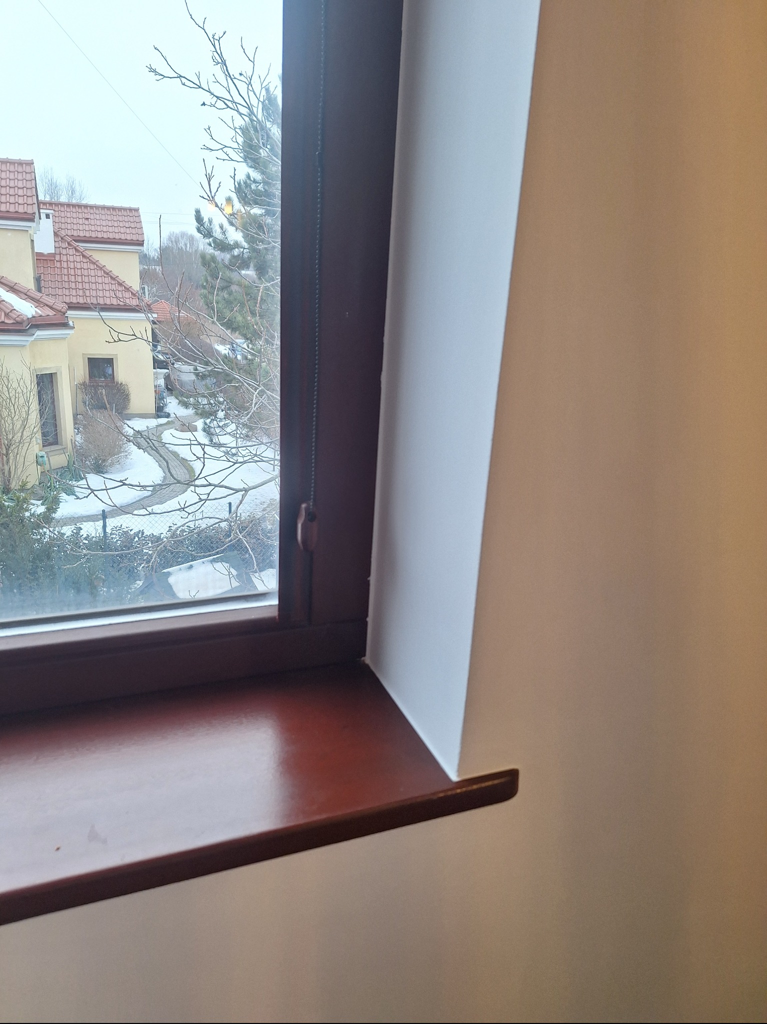 Ciemnobrązowe okno z widokiem na zaśnieżone osiedle domów z czerwonymi dachami. Widoczny fragment białej ściany. Okno z roletą.
