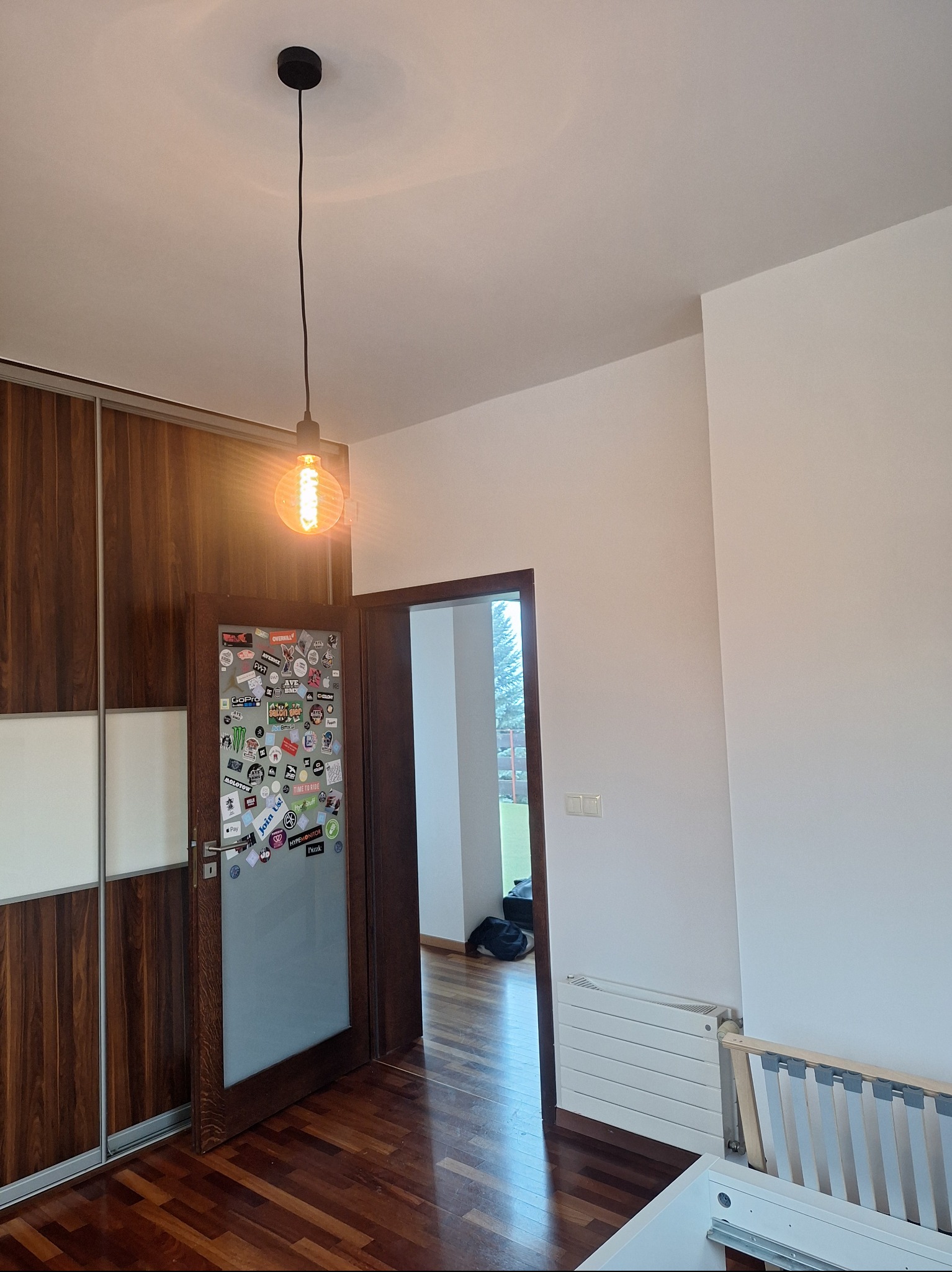 Pokój po remoncie: drewniana podłoga, białe ściany, przesuwne drzwi szafy z drewnianymi panelami, drzwi z naklejkami, wisząca lampa z żarówką Edisona.