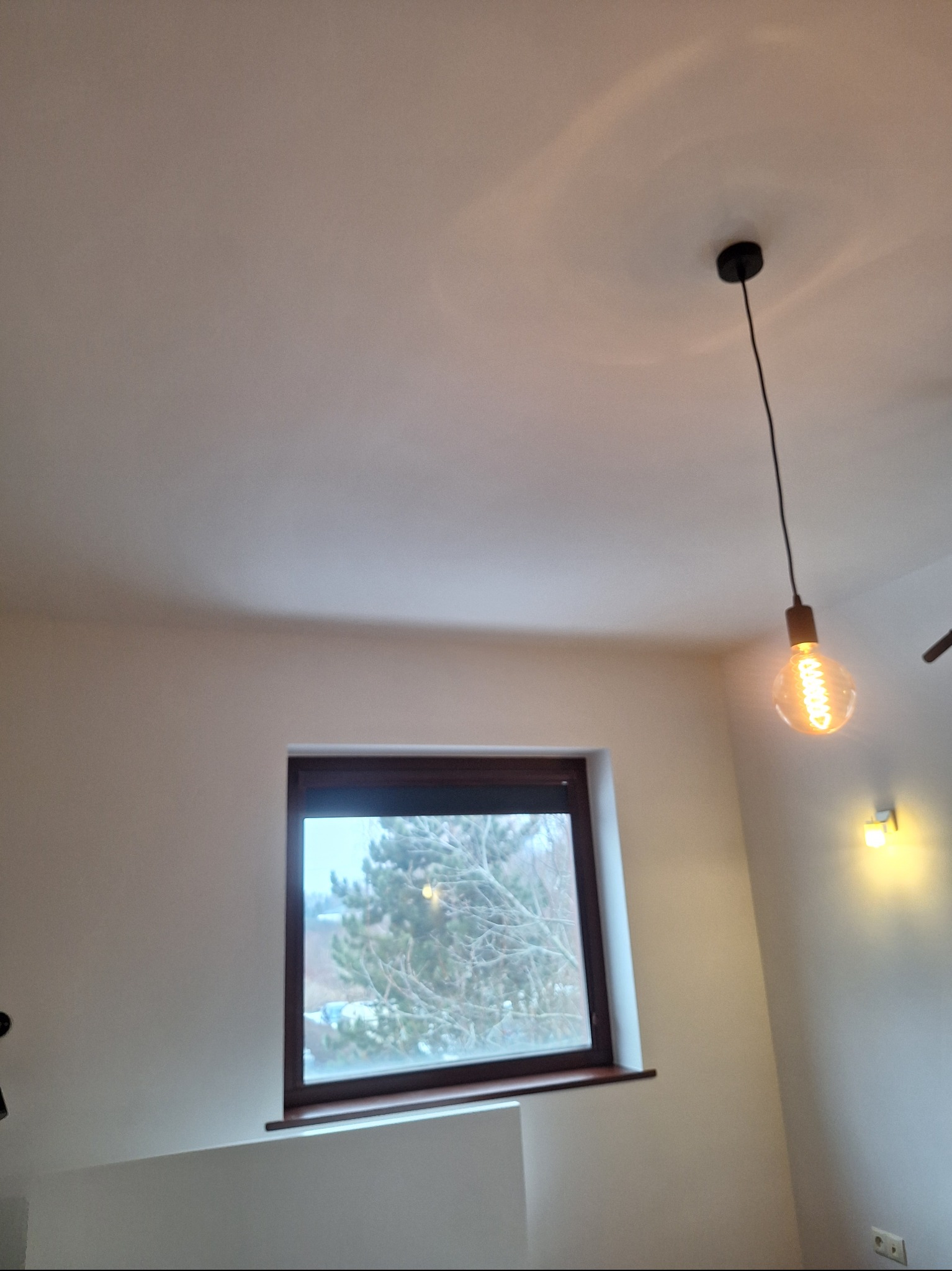 Jasne wnętrze z oknem i wiszącą żarówką edisonowską. Ściany pomalowane na biało, minimalistyczny design. Widok z okna na zewnątrz.