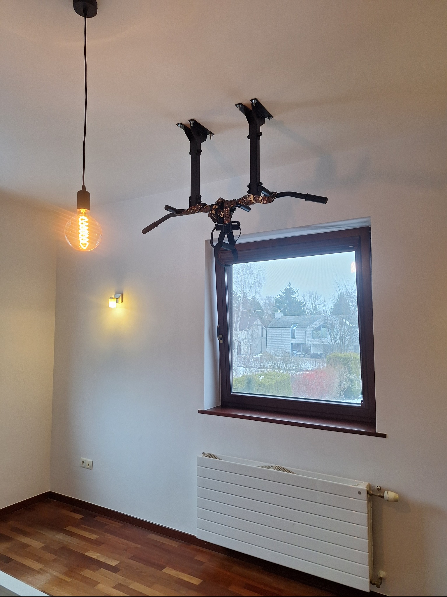 Pokój z zamontowanym drążkiem do podciągania przymocowanym do sufitu, widok na okno i grzejnik. W tle widoczne domy i drzewa.