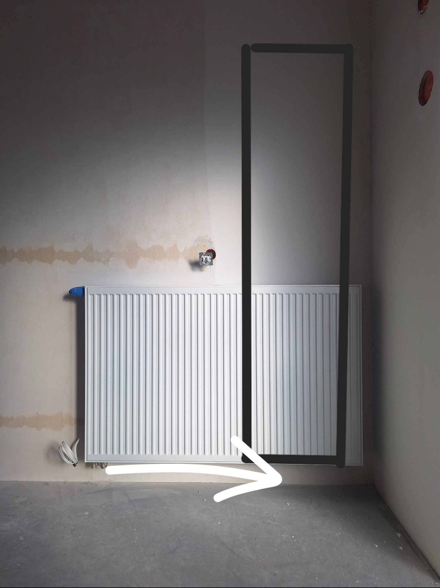 Biały grzejnik na tle surowej ściany z widocznymi śladami po szpachlowaniu i instalacją elektryczną. Zaznaczona rama drzwi i strzałka na podłodze.