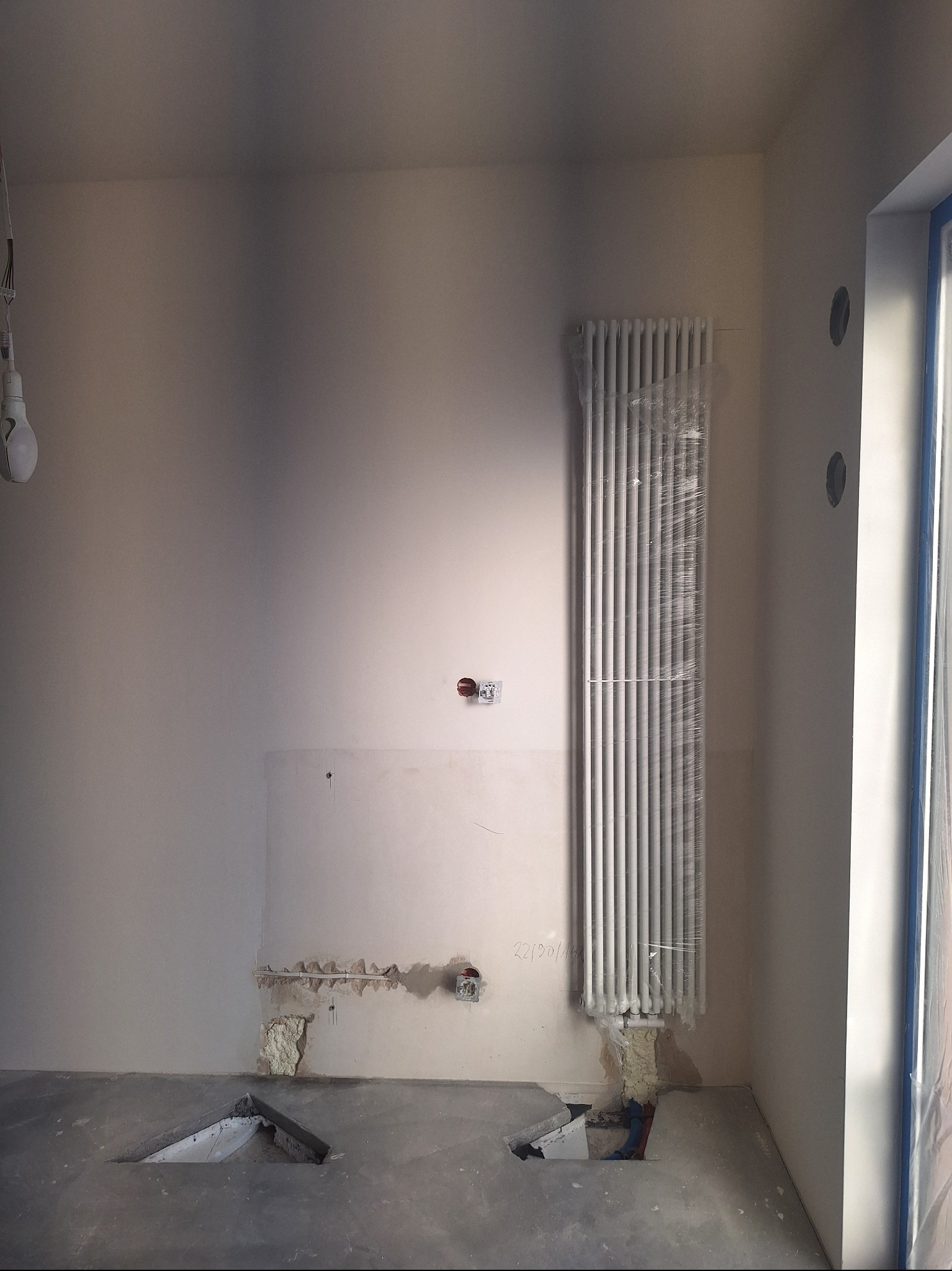 Wnętrze w trakcie prac wykończeniowych: ściany po szpachlowaniu, zabezpieczony grzejnik, otwory w podłodze. Widoczne ślady po instalacjach.