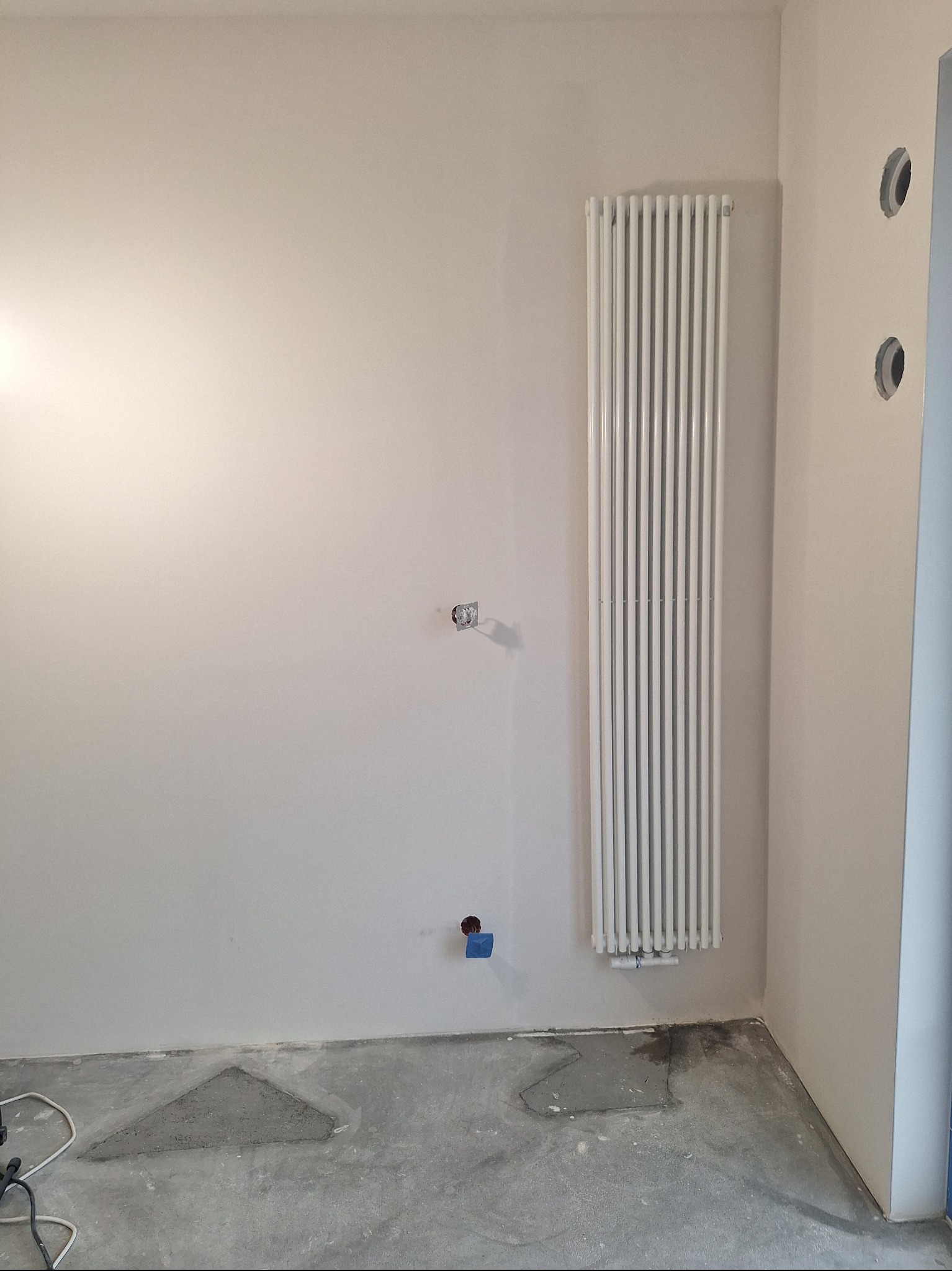 Biały grzejnik panelowy na ścianie w trakcie remontu. Widoczne przygotowane punkty instalacyjne i surowa podłoga. Ściany pomalowane na jasny odcień.
