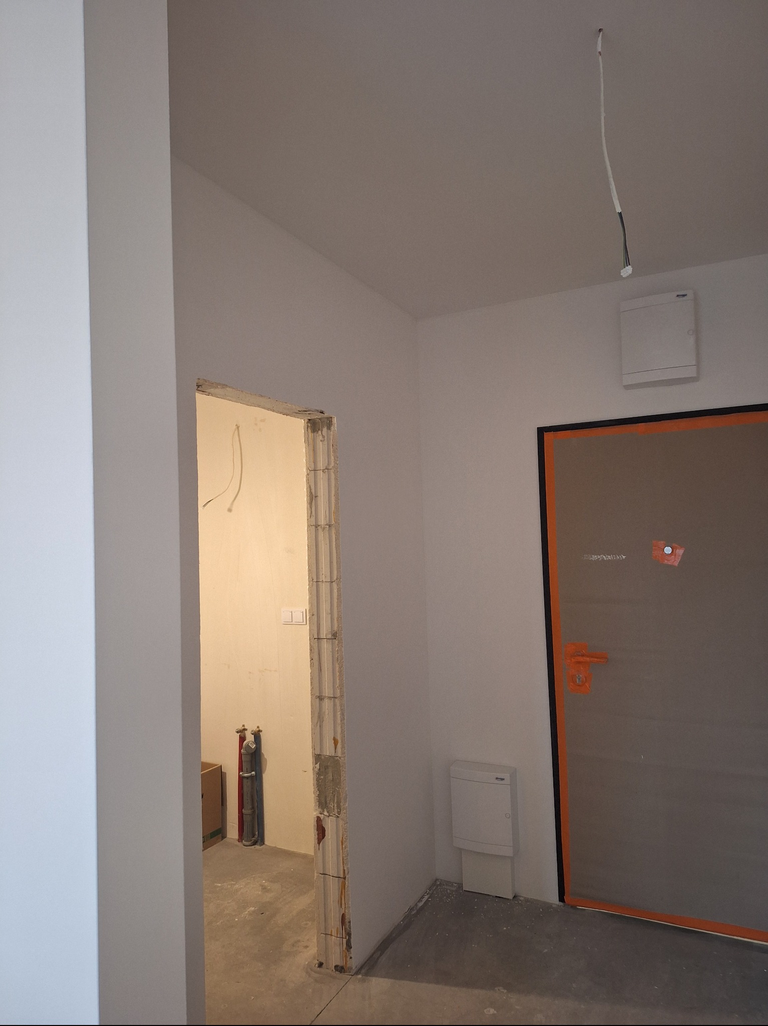 Wnętrze w trakcie remontu: widok na surową ścianę z instalacją elektryczną, ościeżnicę drzwiową zabezpieczoną taśmą i białe ściany. Widoczne elementy instalacji wodnej.