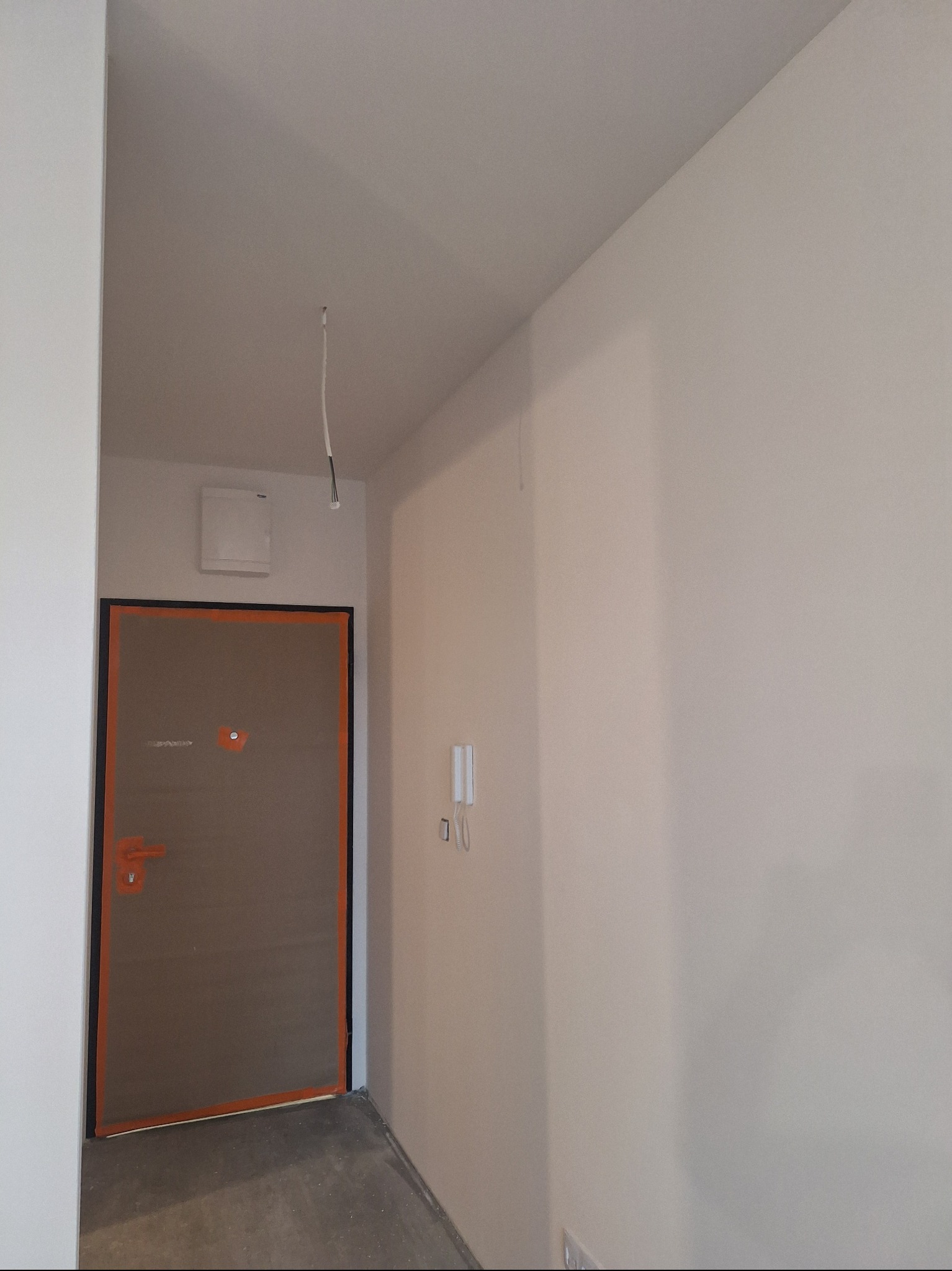 Wnętrze w trakcie prac wykończeniowych: ściany przygotowane do malowania, zabezpieczone drzwi wejściowe, widoczne przewody elektryczne.