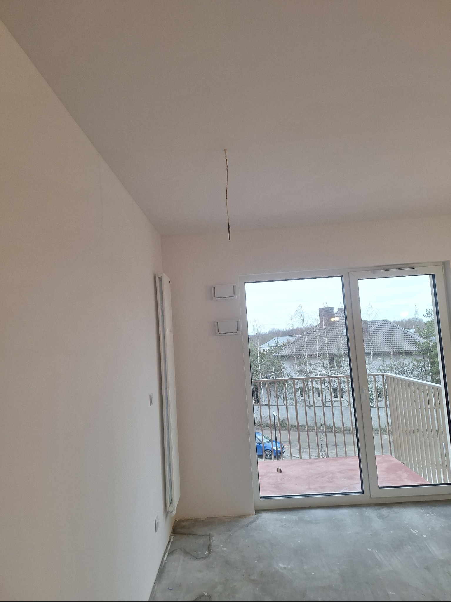 Jasne wnętrze z gładkimi ścianami i widokiem na balkon. Grzejnik na ścianie. Przygotowanie do wykończenia, widoczne niedoskonałości podłogi. Kabel zwisający z sufitu.