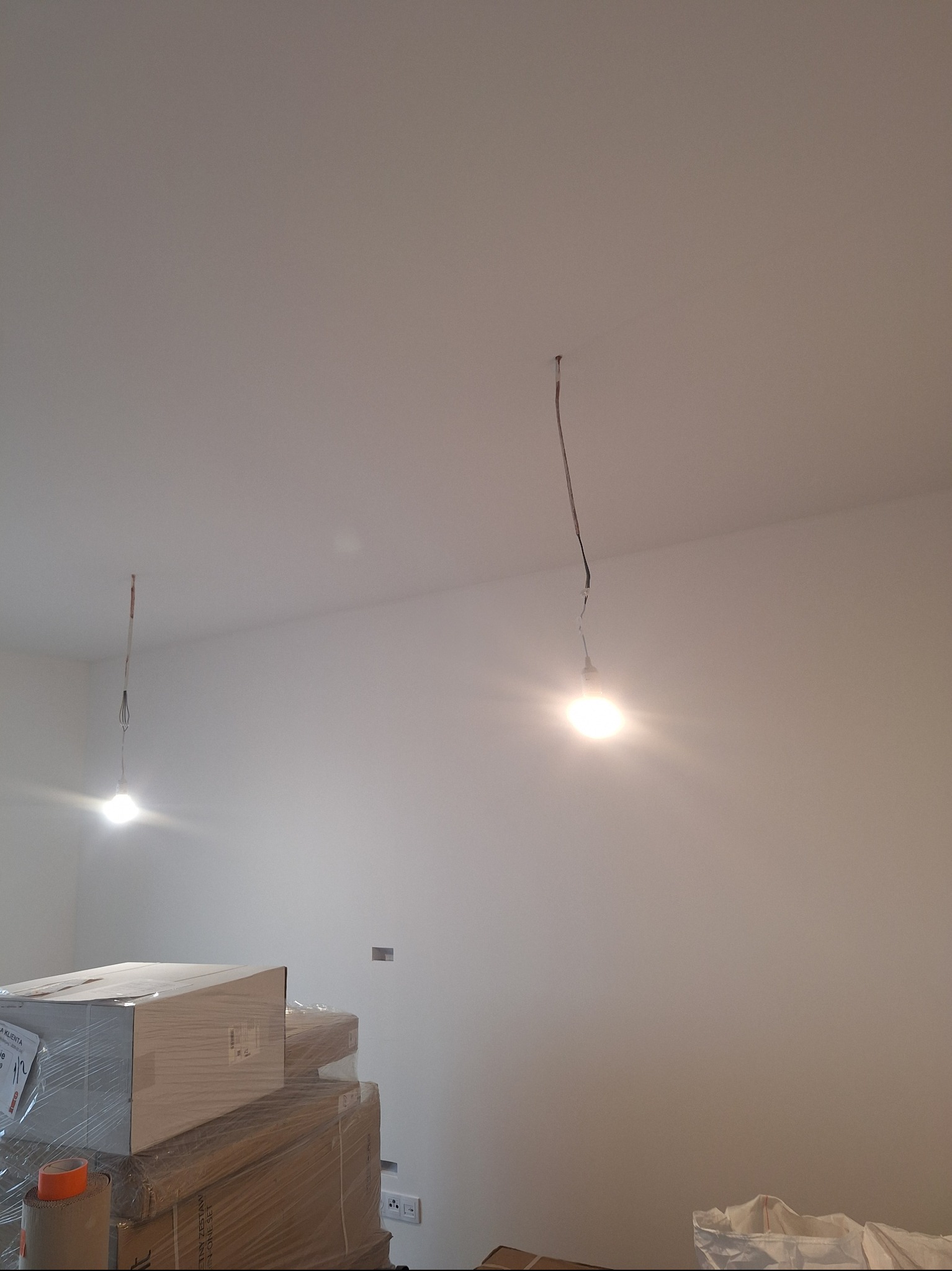 Wnętrze w trakcie prac wykończeniowych: gładkie, białe ściany, wiszące żarówki i zapakowane materiały budowlane. Widoczne gniazdka elektryczne i puszka instalacyjna.