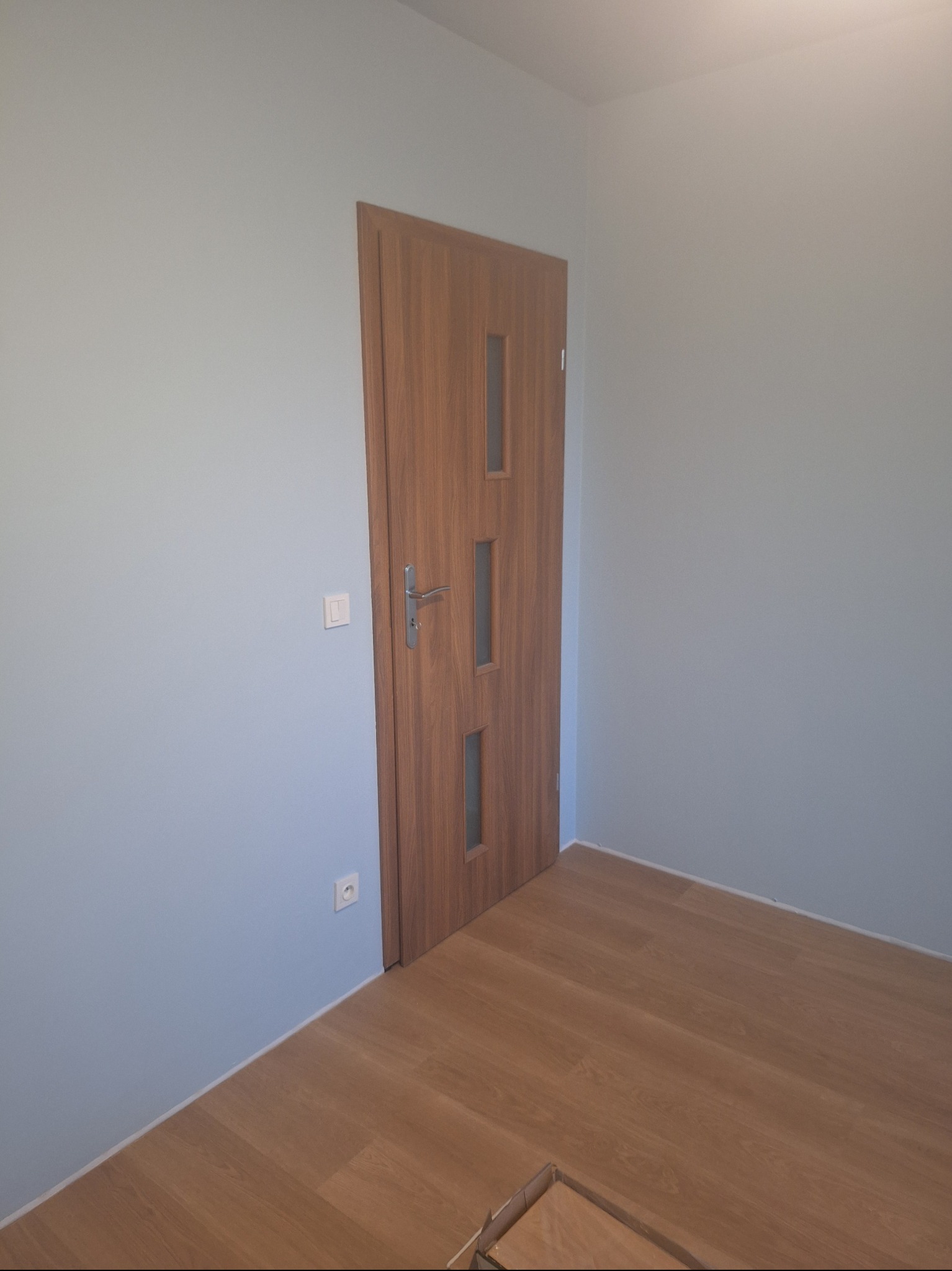 Wnętrze po remoncie: drewniane panele podłogowe, pomalowane na jasny kolor ściany i zamontowane drzwi z przeszkleniami. Widoczne gniazdka i włącznik.