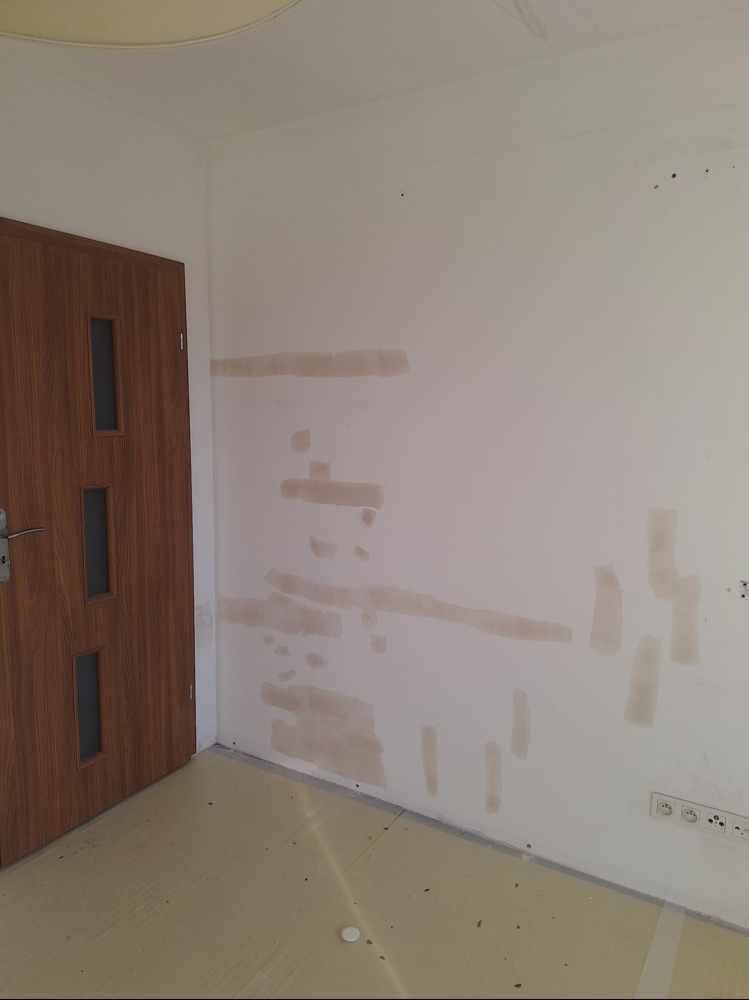 Przygotowana ściana do szpachlowania z widocznymi łatami, brązowe drzwi z przeszkleniami po lewej stronie, podłoga zabezpieczona folią malarską.