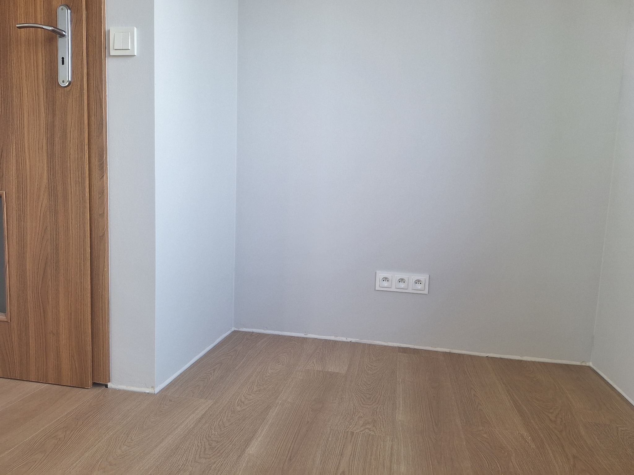Pomalowane na szaro wnętrze pokoju z drewnianą podłogą i drzwiami. Widoczne gniazdka elektryczne i włącznik światła. Minimalistyczny design.