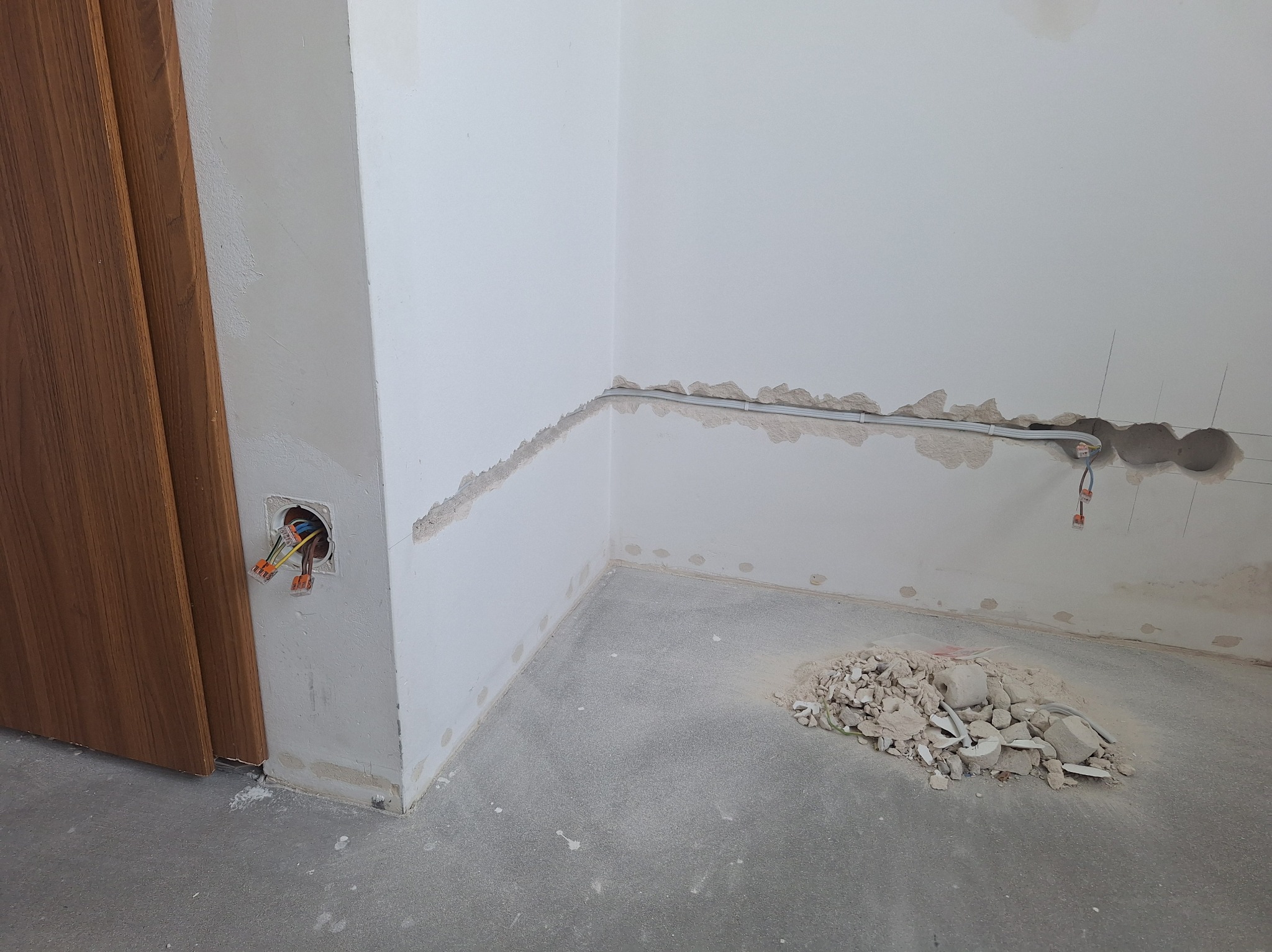 Wnętrze w trakcie prac przygotowawczych: ściany z widocznymi śladami po kuciu, wystające przewody elektryczne, gruz na podłodze, fragment ościeżnicy drzwiowej.