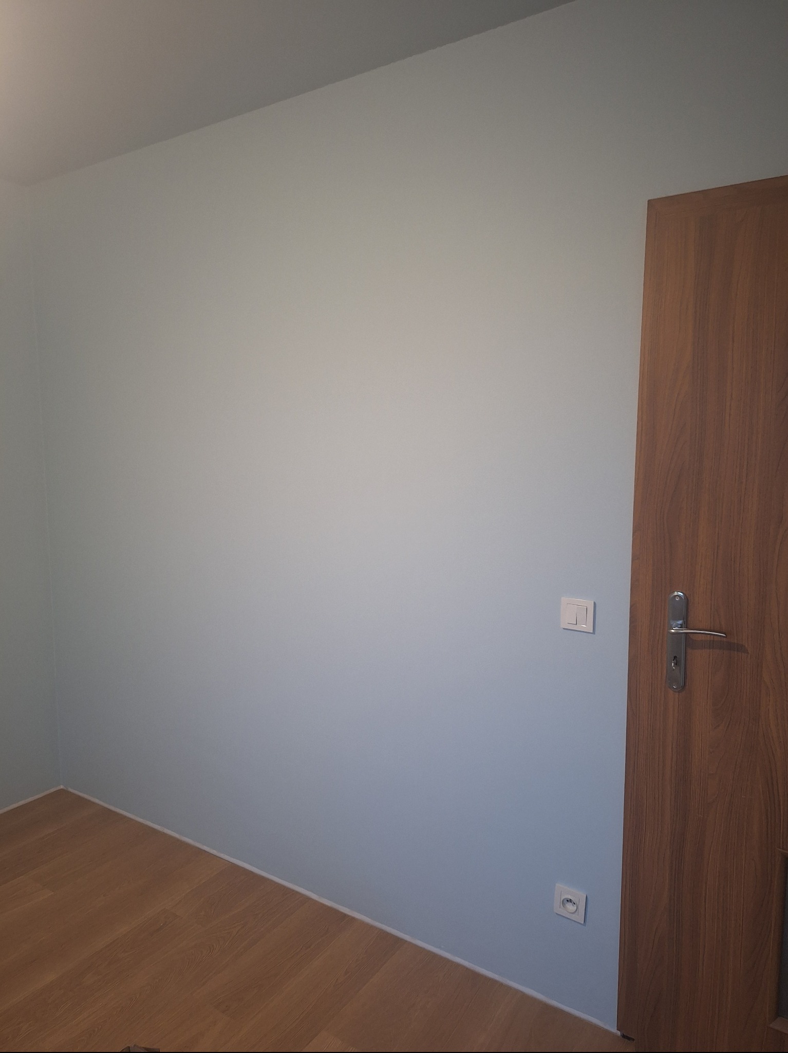 Pomalowane na jasny odcień błękitu ściany pokoju z drewnianą podłogą i drzwiami. Widoczny włącznik światła i gniazdko elektryczne.