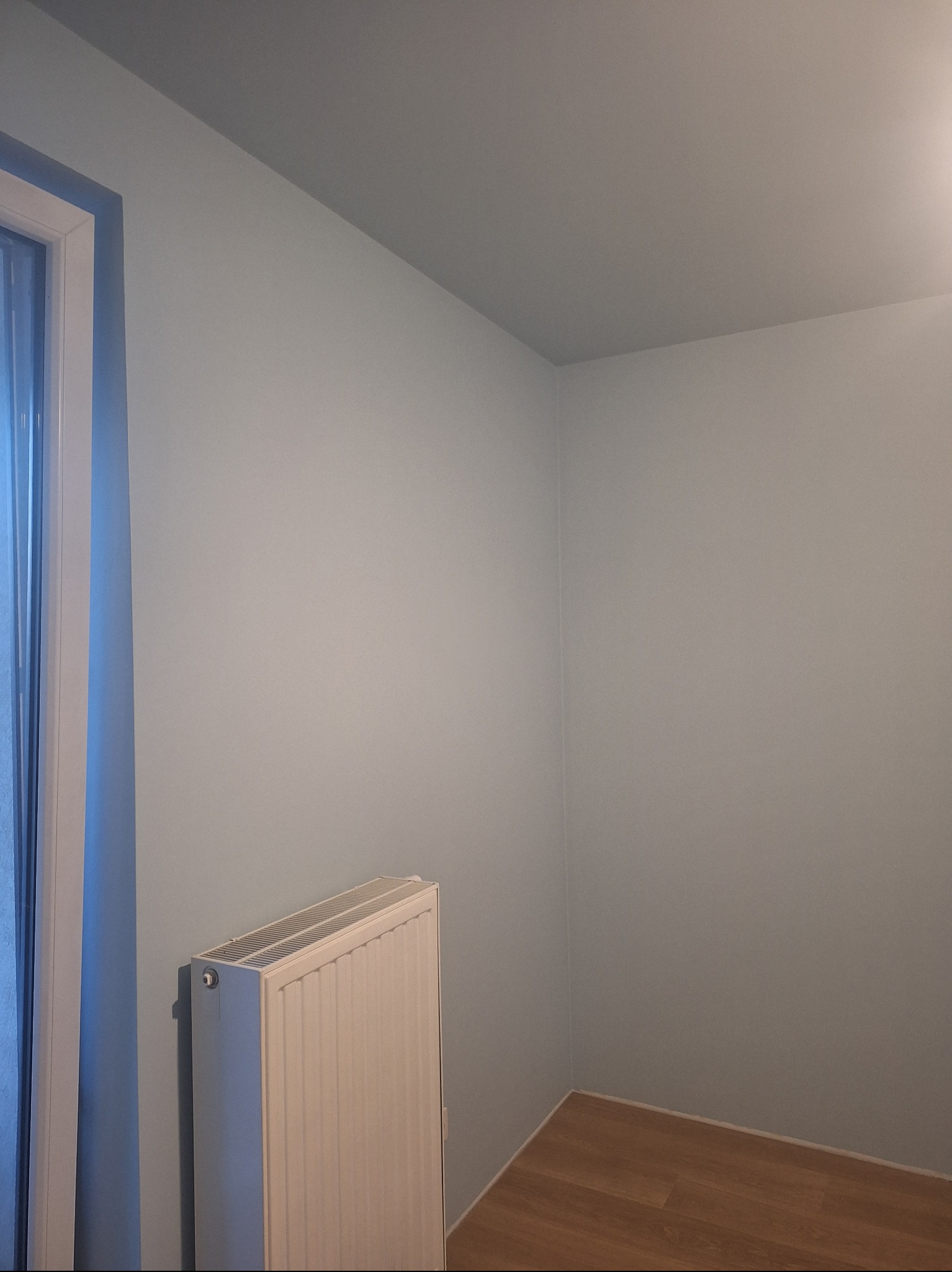 Jasny pokój po malowaniu ścian na pastelowy odcień błękitu, z widocznym grzejnikiem i fragmentem podłogi z paneli. Świeżo pomalowane ściany.