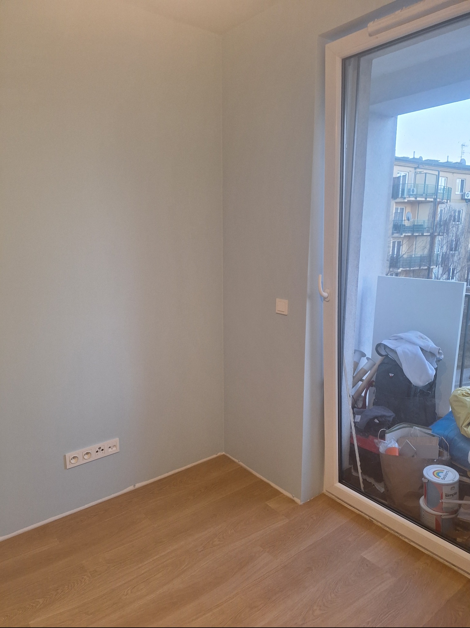 Jasne, świeżo pomalowane wnętrze z drewnianą podłogą i widokiem na balkon przez duże okno. Widoczne gniazdka elektryczne i puszka farby.