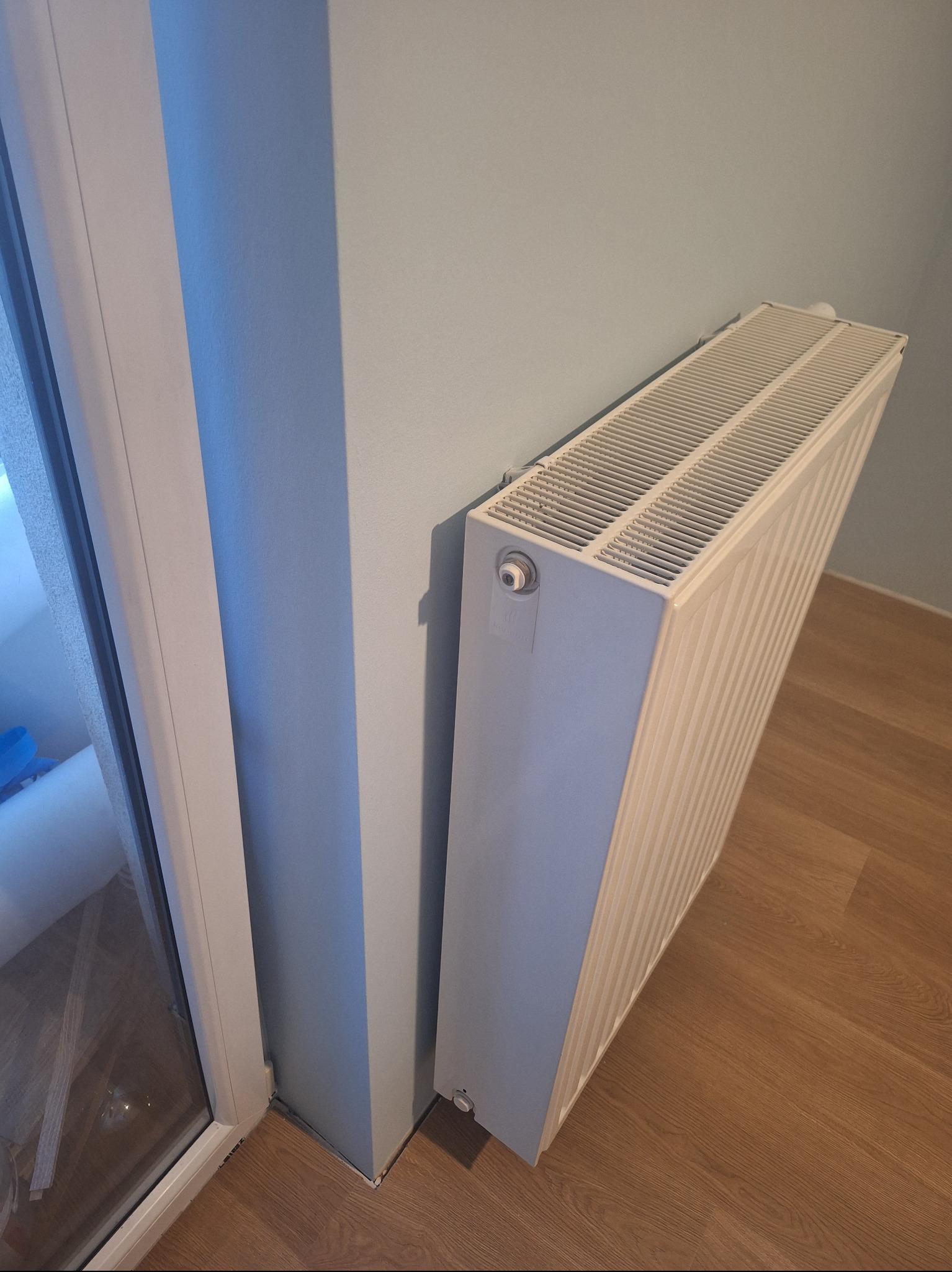 Biały grzejnik panelowy zamontowany przy pomalowanej na jasny błękit ścianie, obok białej ramy okiennej i drewnianej podłogi. Ujęcie z dołu.