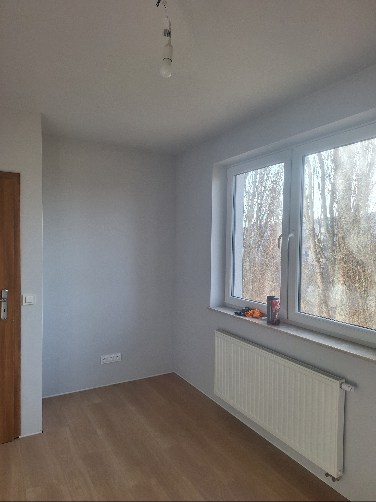 Jasne wnętrze po odświeżeniu: świeżo pomalowane ściany w odcieniach szarości, panele podłogowe, biały grzejnik pod oknem z widokiem na drzewa.