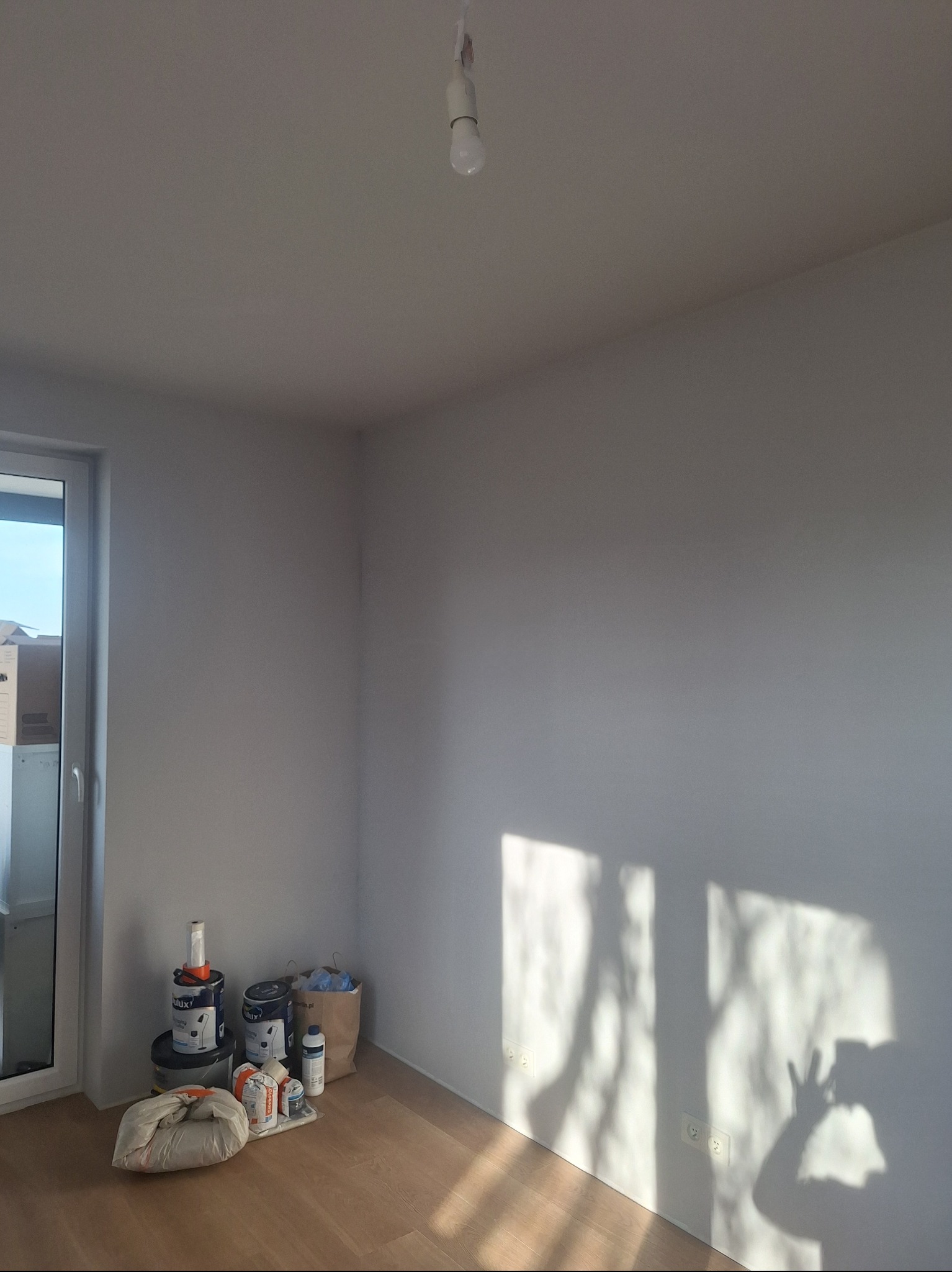 Pomalowane na biało wnętrze pokoju z widocznymi puszkami farb i workiem gładzi przy ścianie. Na ścianie cień okna i osoby robiącej zdjęcie.