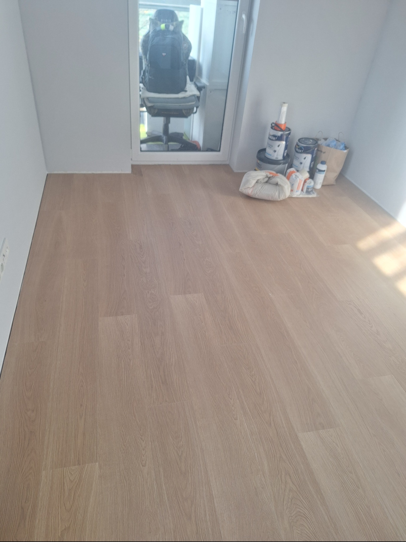Jasne panele podłogowe ułożone w pokoju, widoczne puszki z farbą i akcesoria malarskie przy ścianie. W tle krzesło biurowe przy oknie.