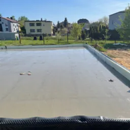 Świeżo wylany fundament domu jednorodzinnego w słoneczny dzień, z widoczną izolacją obwodową i elementami instalacji w posadzce.