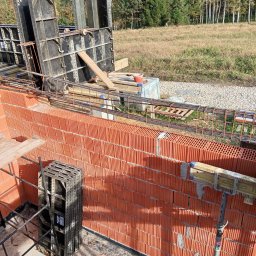 R.D. - Bud Budowa - Remonty RAFAŁ SKATUŁA - Wznoszenie muru z czerwonej cegły zbrojonego stalą, z użyciem szalunków systemowych. Widoczne elementy konstrukcyjne i narzędzia na tle krajobrazu.