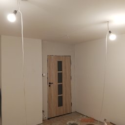 R.D. - Bud Budowa - Remonty RAFAŁ SKATUŁA - Wnętrze pokoju w trakcie odświeżania: białe ściany, drewniane drzwi z szybami, prowizoryczne oświetlenie i wiadra z farbą na podłodze. Widoczne ślady prac malarskich.