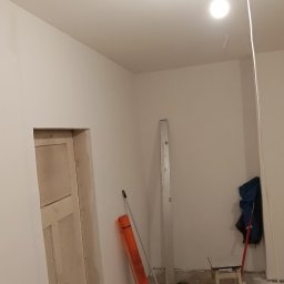 R.D. - Bud Budowa - Remonty RAFAŁ SKATUŁA - Wnętrze w trakcie odświeżania: białe ściany, surowe wykończenie ościeżnicy, narzędzia malarskie i prowizoryczne oświetlenie. Widok na remontowany pokój.