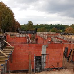 R.D. - Bud Budowa - Remonty RAFAŁ SKATUŁA - Ceglane ściany fundamentowe z drewnianym szalunkiem na tle pochmurnego nieba i drzew. Widoczne zbrojenia i elementy konstrukcyjne.
