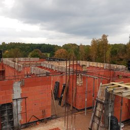 R.D. - Bud Budowa - Remonty RAFAŁ SKATUŁA - Wysoki kąt: Czerwone bloczki ścian domu w trakcie budowy, z drewnianym szalunkiem i zbrojeniami, na tle lasu pod pochmurnym niebem. Widoczne narzędzia i materiały budowlane.