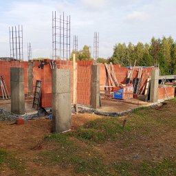 R.D. - Bud Budowa - Remonty RAFAŁ SKATUŁA - Budowa domu: ściany z czerwonej cegły, betonowe słupy i zbrojenia na tle zieleni i błękitnego nieba. Widoczne materiały budowlane i elementy konstrukcyjne.