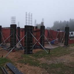 R.D. - Bud Budowa - Remonty RAFAŁ SKATUŁA - Wznoszenie konstrukcji domu: zbrojenie kolumn w szalunkach, widoczne ceglane ściany w tle, pochmurna pogoda. Elementy konstrukcyjne na placu budowy.