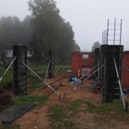 R.D. - Bud Budowa - Remonty RAFAŁ SKATUŁA - Budowa domu w trakcie: widoczne szalunki kolumnowe ze zbrojeniem, fragment muru z czerwonej cegły. Mglista pogoda, drzewa w tle. Ujęcie z poziomu gruntu.
