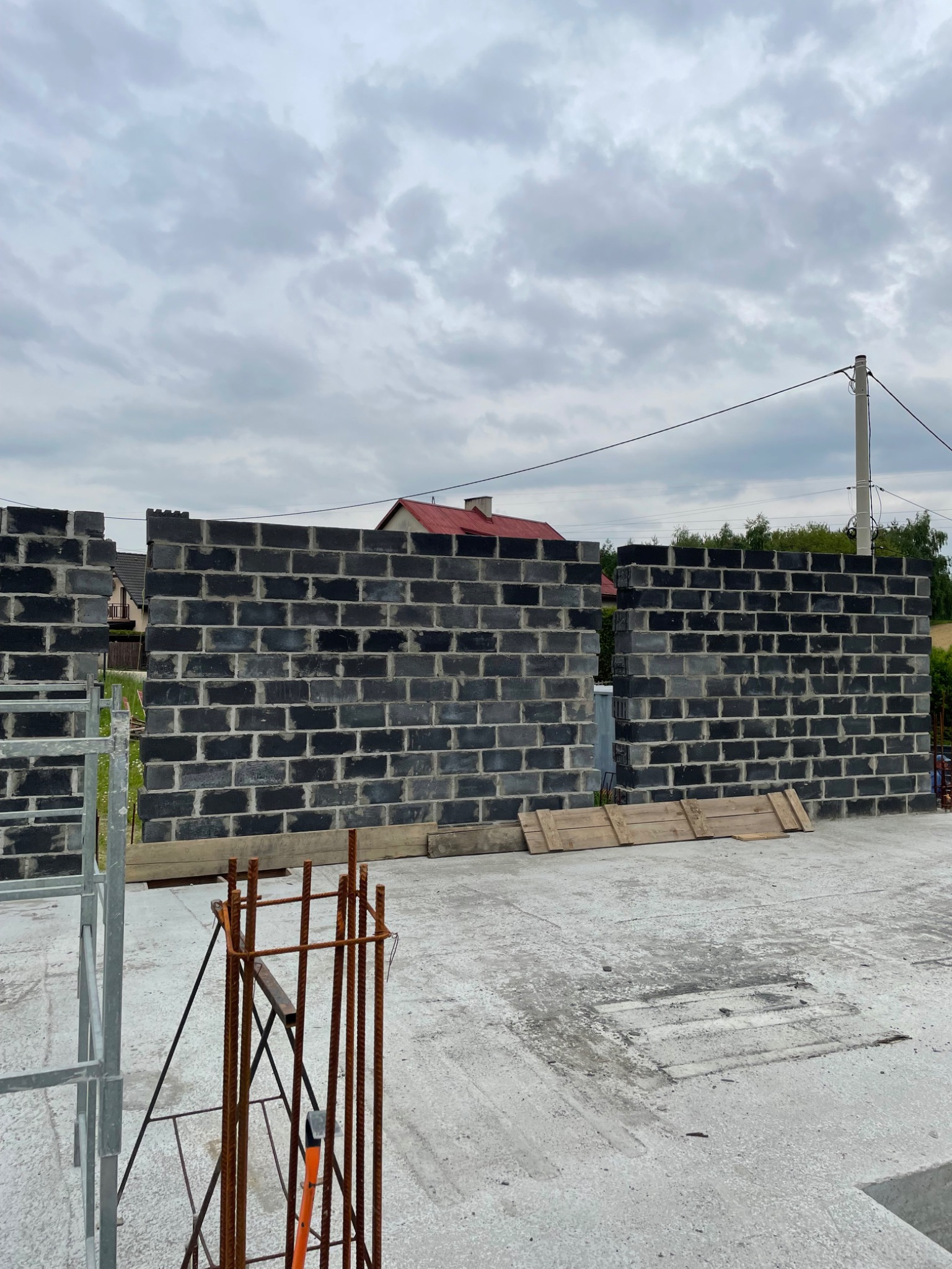 Wznoszenie ścian z szarego bloczka betonowego na fundamencie. Widoczne zbrojenia i elementy szalunkowe. Tło: pochmurne niebo i fragment zabudowy jednorodzinnej.