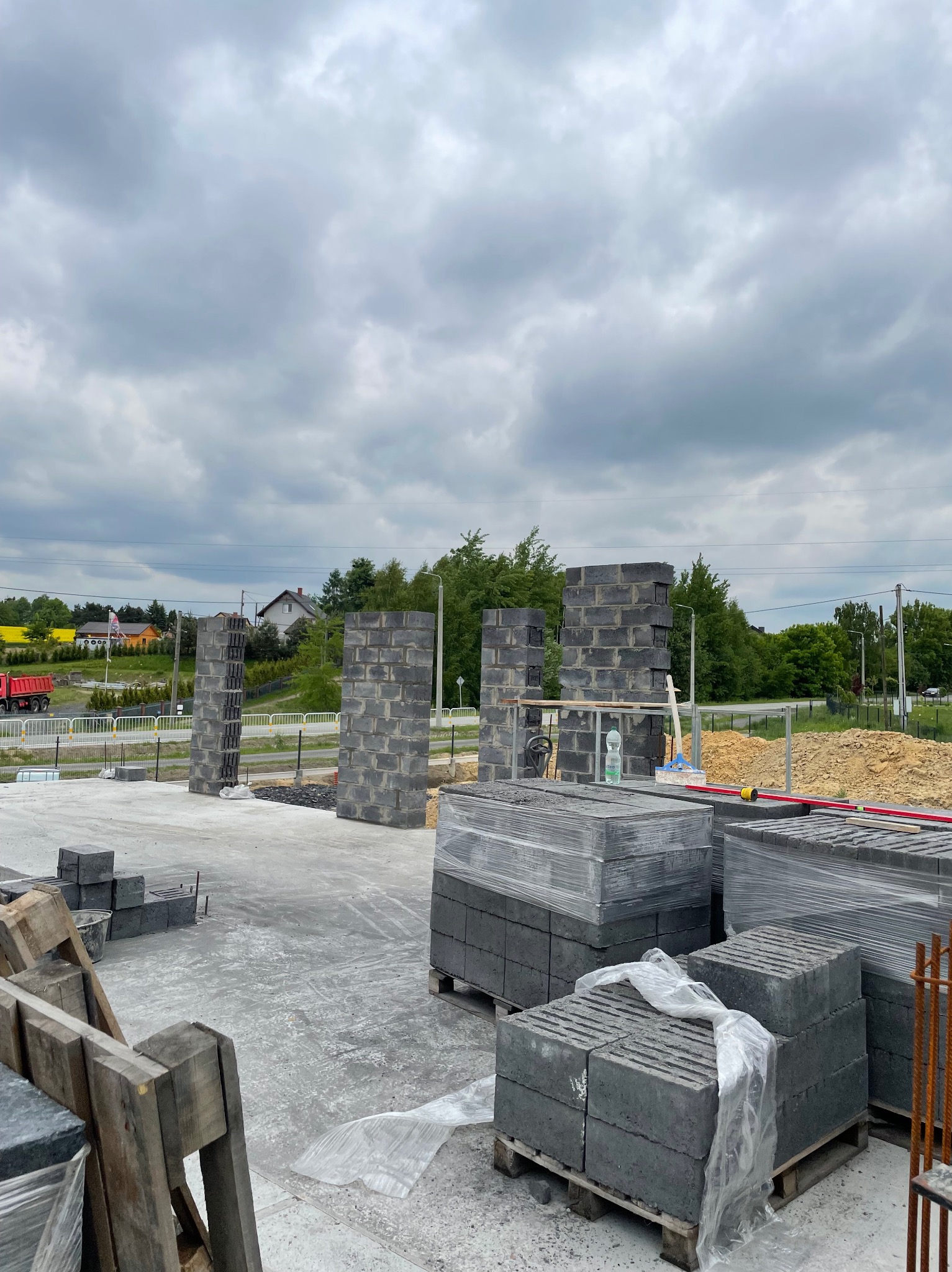 Wznoszone ściany z bloczków betonowych na fundamencie. Palety z bloczkami czekają na użycie. Widok na plac budowy w pochmurny dzień. Częściowo wzniesione ściany.