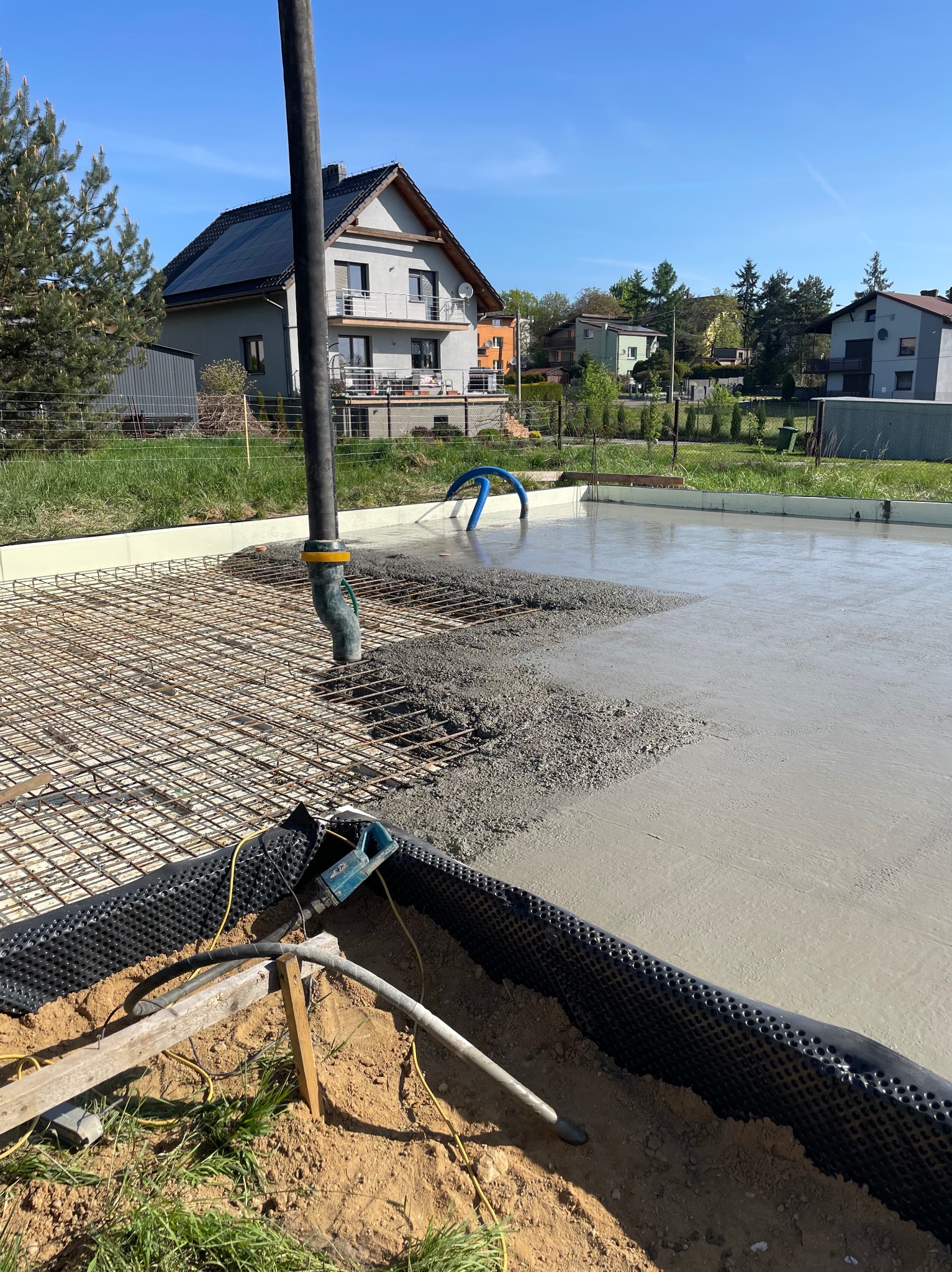 Wylewka betonu na zbrojenie fundamentu domu jednorodzinnego w słoneczny dzień. Widoczna pompa do betonu i częściowo zalana powierzchnia. W tle dom z panelami słonecznymi.