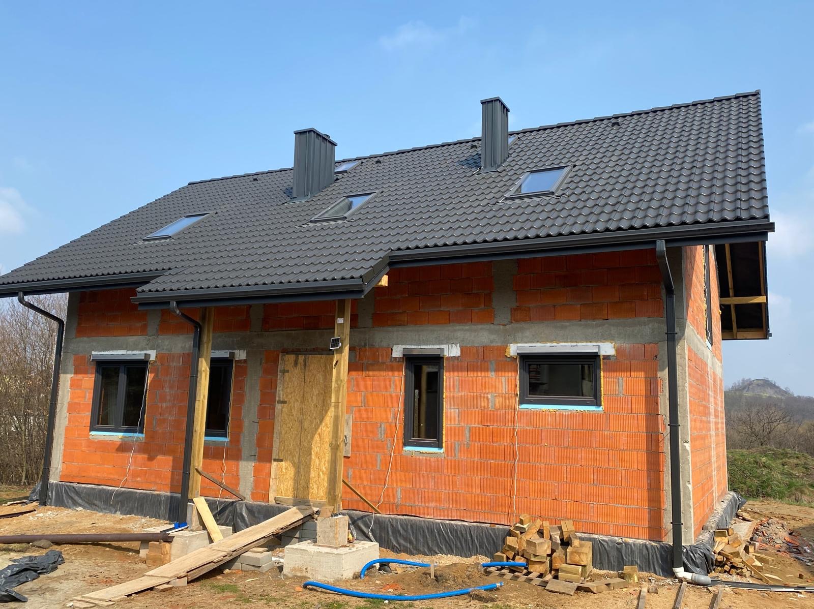 Surowy dom z czerwonej cegły z grafitową dachówką i oknami. Widoczne kominy, rynny, okna dachowe. Częściowo ocieplone ściany. W tle wzgórze i błękitne niebo.