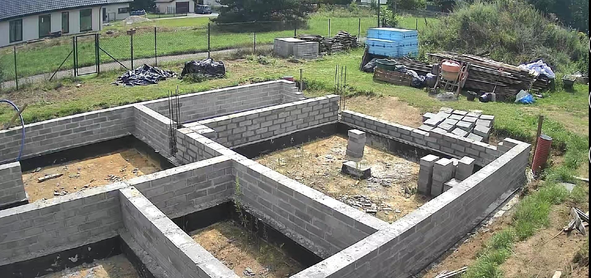 Wysoki kąt na fundamenty z szarych bloczków betonowych, częściowo zalane czarną izolacją, z wystającymi prętami zbrojeniowymi. Na trawie składowane materiały budowlane.