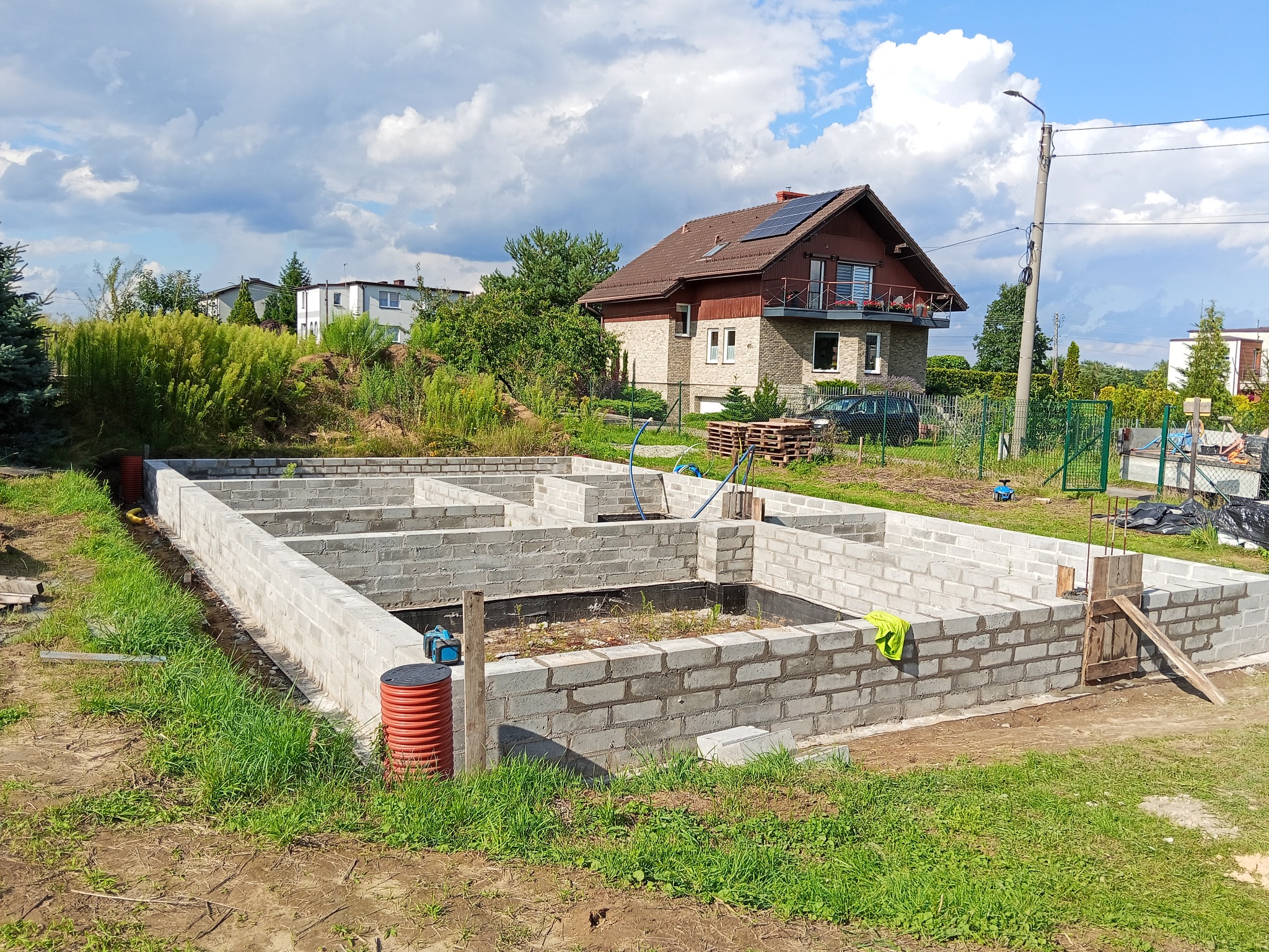 Wykopane i wymurowane fundamenty domu z bloczków betonowych, widoczne izolacje pionowe, w tle dom jednorodzinny i błękitne niebo z chmurami.