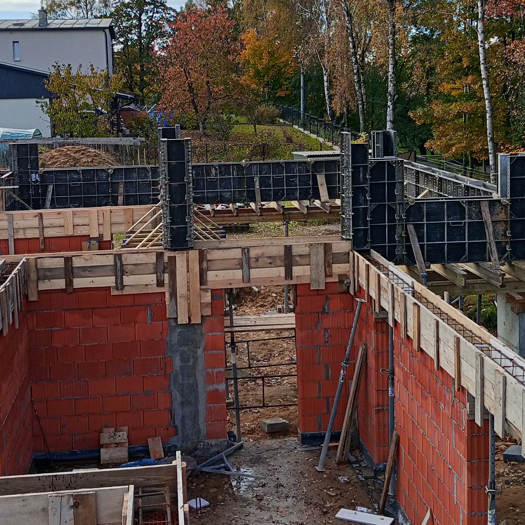 Wznoszenie ścian z czerwonej cegły z drewnianym szalunkiem i metalowymi stemplami. Widok z góry na plac budowy w jesiennej scenerii.