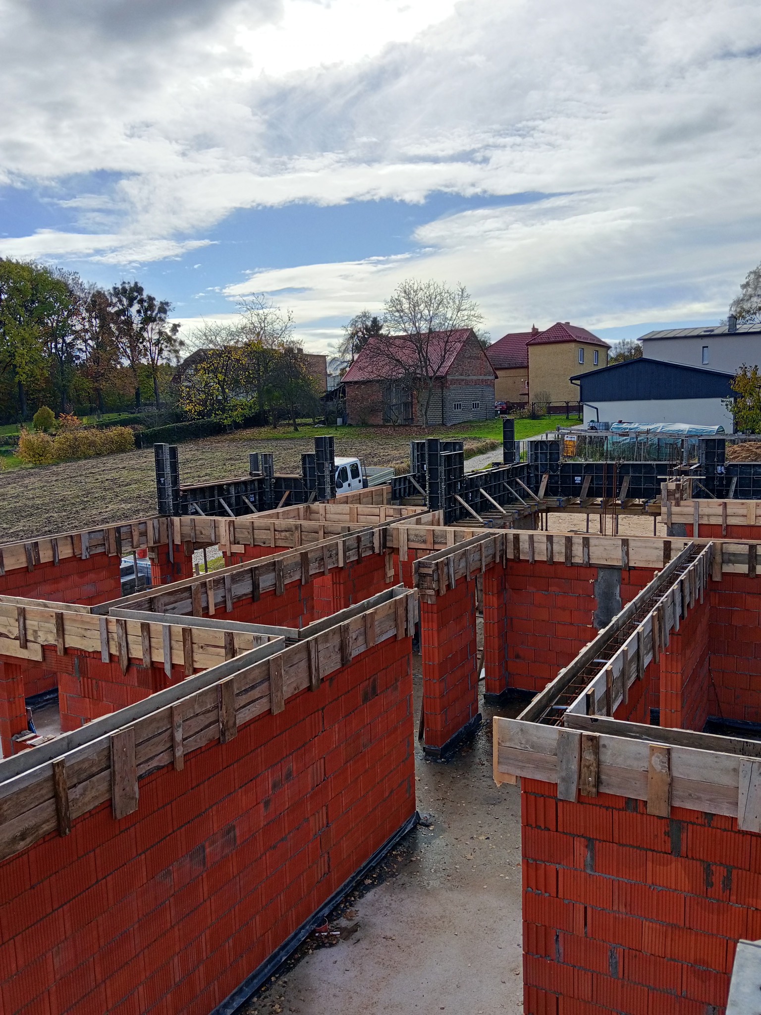 Czerwone ściany z pustaków ceramicznych w trakcie budowy domu, z widocznym szalunkiem i zbrojeniem, na tle zabudowy jednorodzinnej i pochmurnego nieba.