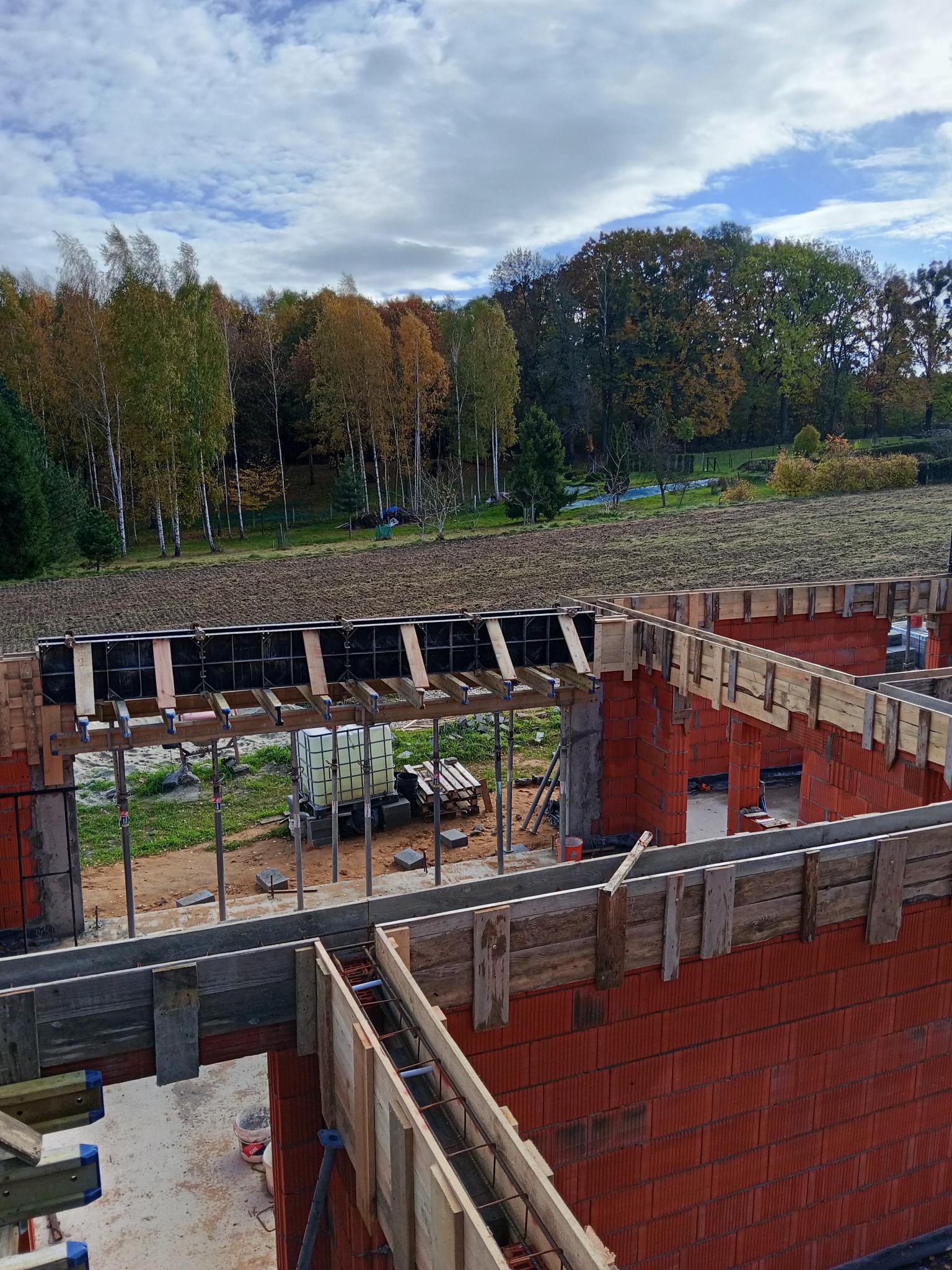 Wznoszenie ścian domu z czerwonej cegły, widoczne szalunki i zbrojenia. W tle drzewa z jesiennymi liśćmi i błękitne niebo z chmurami. Budowa w toku, widok z góry.