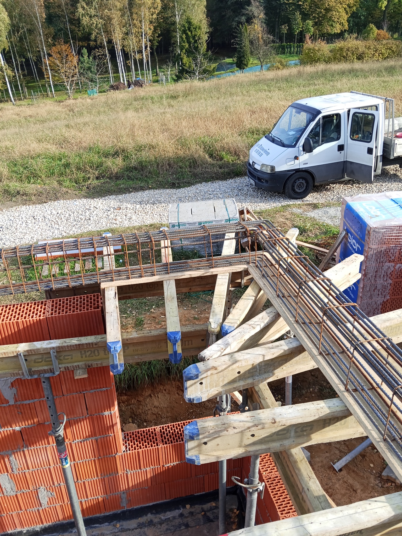 Wysoki kąt: Zbrojenie fundamentów z ceglanymi ścianami, drewnianym szalunkiem i dostawczym busem w tle, na tle zieleni i drzew w słoneczny dzień.