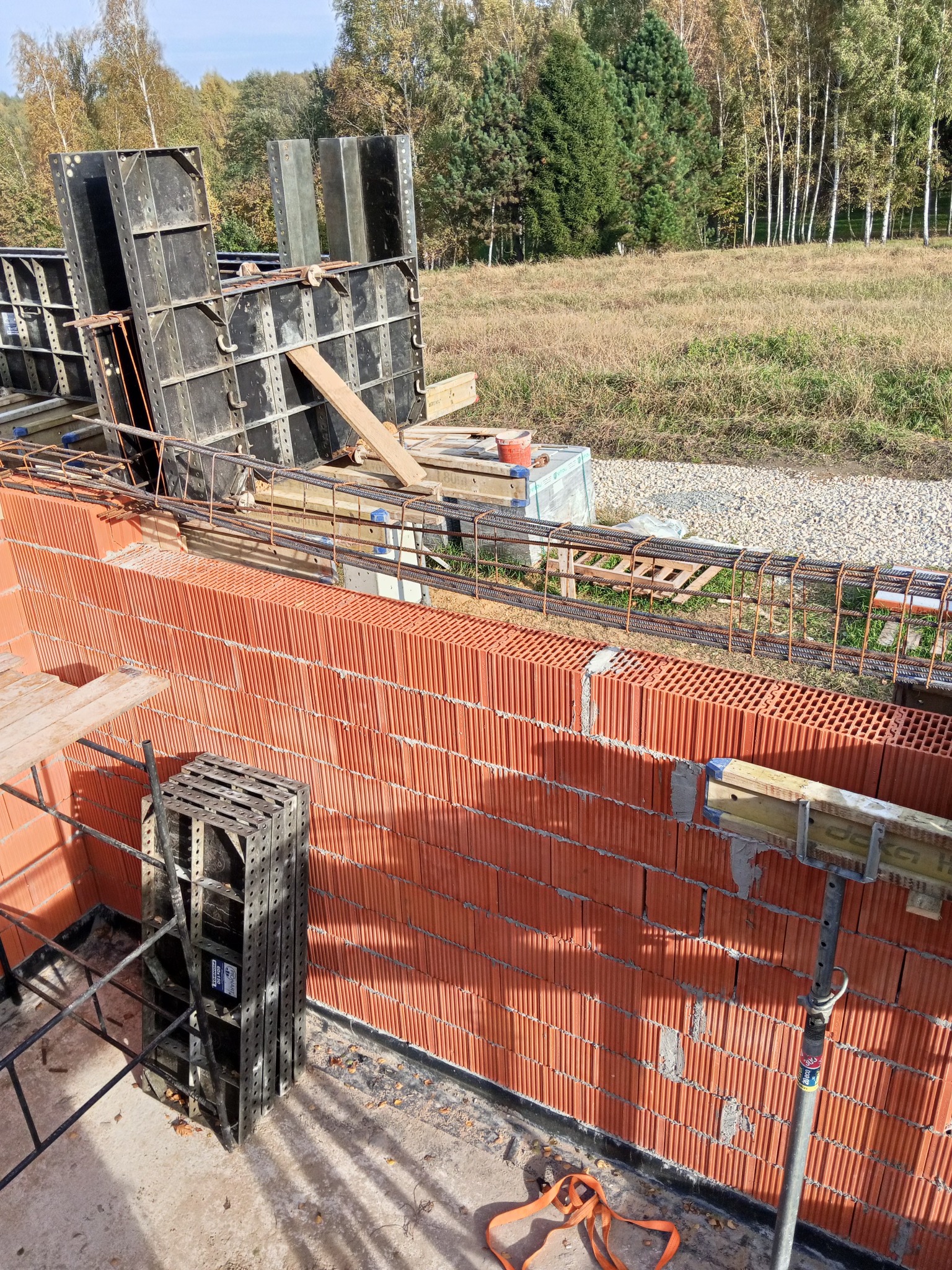 Wznoszenie muru z czerwonej cegły zbrojonego stalą, z użyciem szalunków systemowych. Widoczne elementy konstrukcyjne i narzędzia na tle krajobrazu.