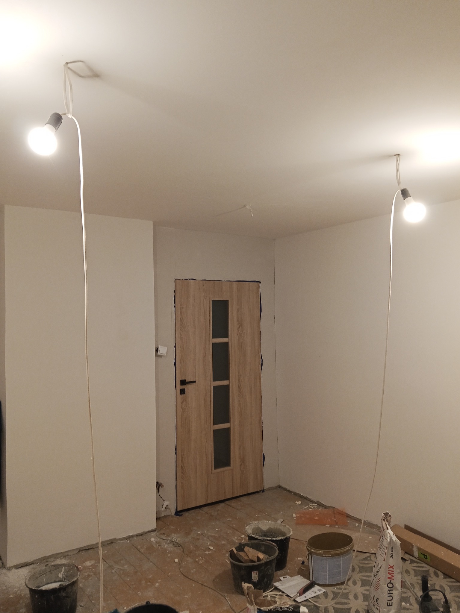 Wnętrze pokoju w trakcie odświeżania: białe ściany, drewniane drzwi z szybami, prowizoryczne oświetlenie i wiadra z farbą na podłodze. Widoczne ślady prac malarskich.