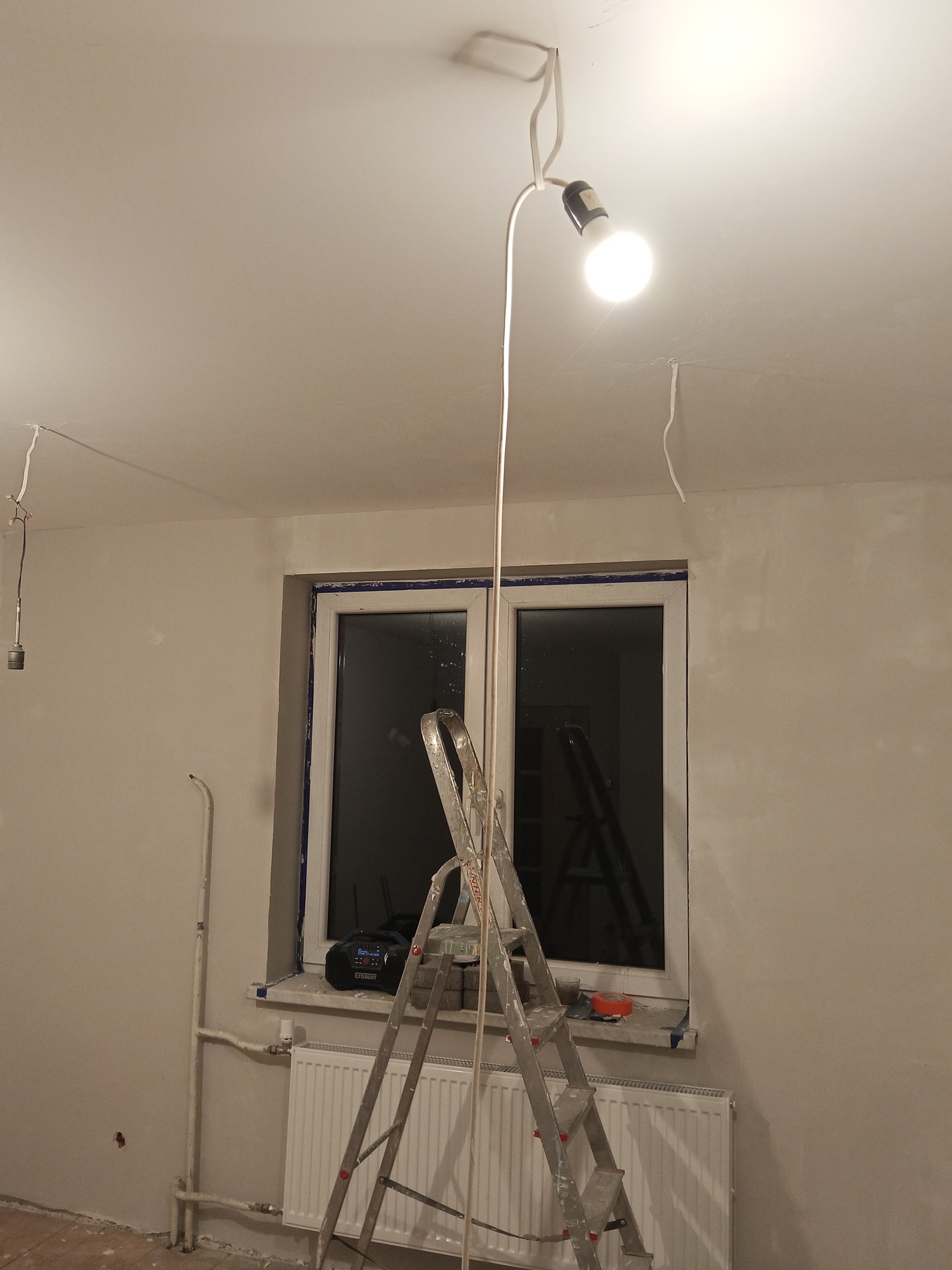 Wnętrze w trakcie remontu: drabina oparta o okno, widoczne przewody elektryczne, ściany po szpachlowaniu, tymczasowe oświetlenie. Etap przygotowania do malowania.