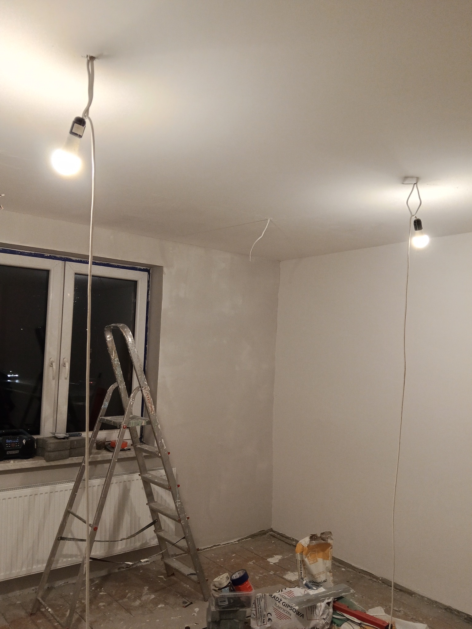 Pokój w trakcie prac wykończeniowych: drabina, gładzie gipsowe, prowizoryczne oświetlenie. Ściany częściowo pokryte gładzią, widoczne narzędzia i materiały budowlane.