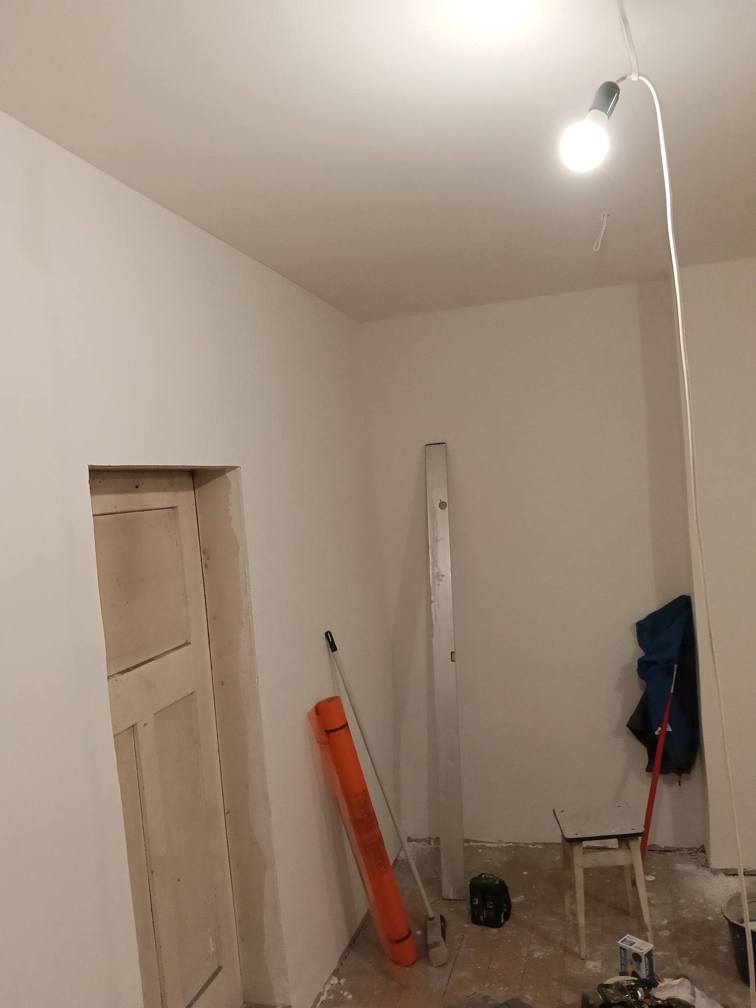 Wnętrze w trakcie odświeżania: białe ściany, surowe wykończenie ościeżnicy, narzędzia malarskie i prowizoryczne oświetlenie. Widok na remontowany pokój.