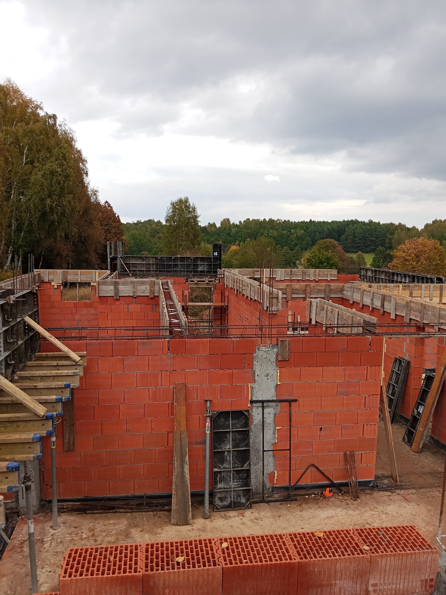 Ceglane ściany fundamentowe z drewnianym szalunkiem na tle pochmurnego nieba i drzew. Widoczne zbrojenia i elementy konstrukcyjne.
