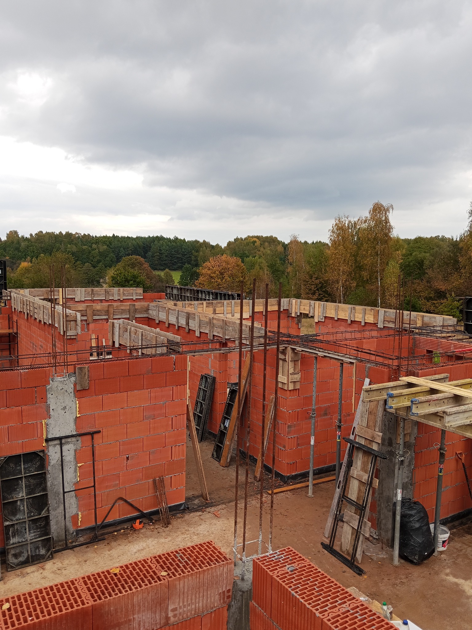 Wysoki kąt: Czerwone bloczki ścian domu w trakcie budowy, z drewnianym szalunkiem i zbrojeniami, na tle lasu pod pochmurnym niebem. Widoczne narzędzia i materiały budowlane.
