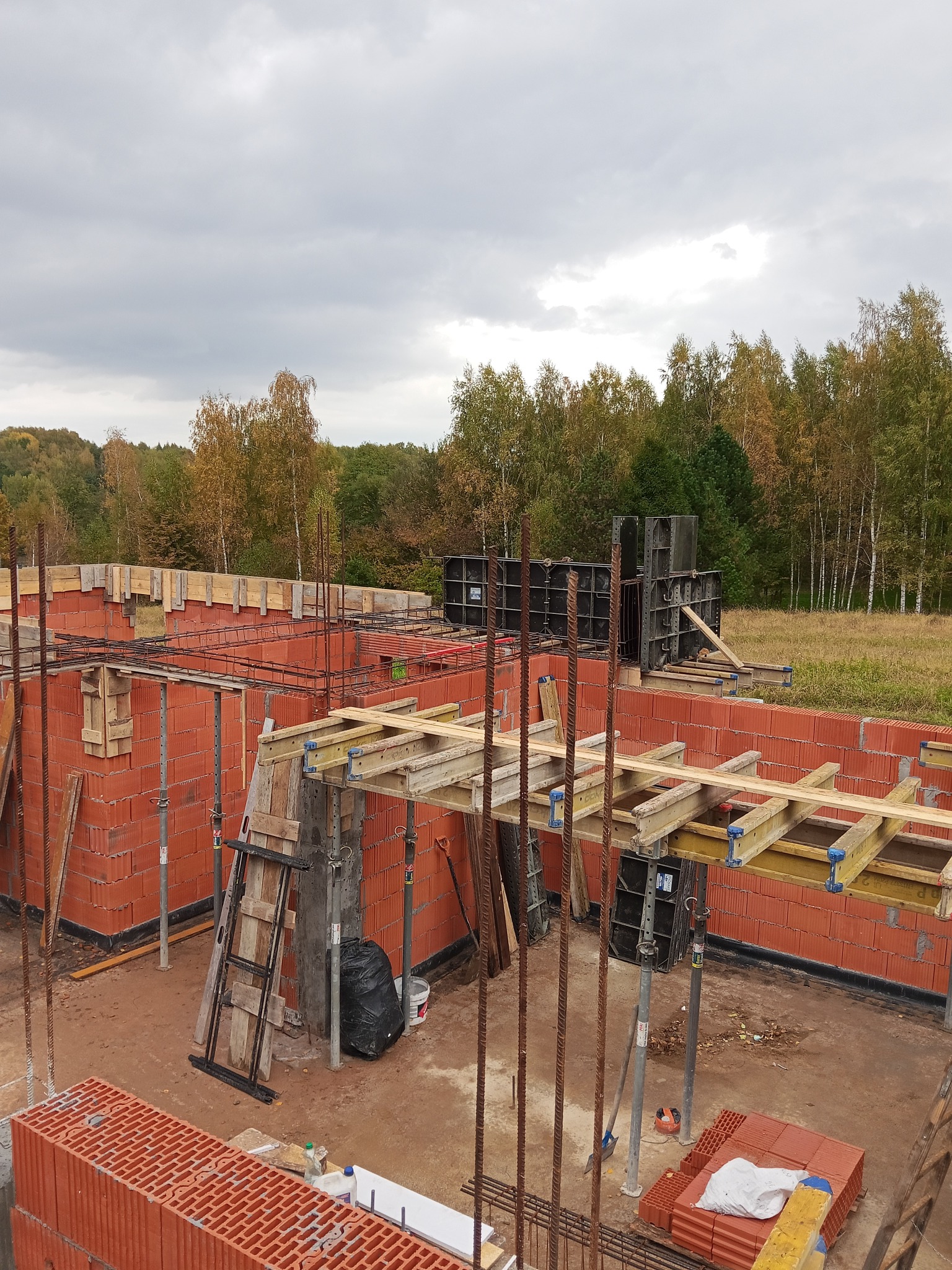 Budowa domu z czerwonej cegły: ściany wzniesione, widoczne zbrojenia i szalunki. W tle drzewa i pochmurne niebo. Etap prac murarskich.